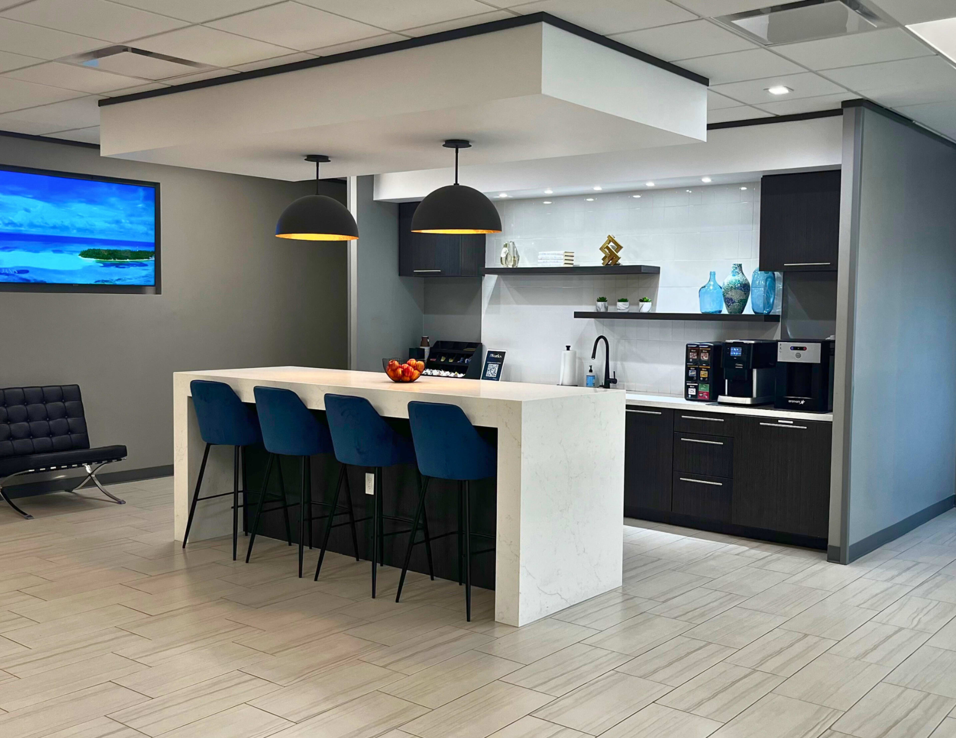 The image shows a modern kitchen area with a marble-topped island and four blue bar stools, complemented by dark cabinetry and appliances, along with a large TV display.