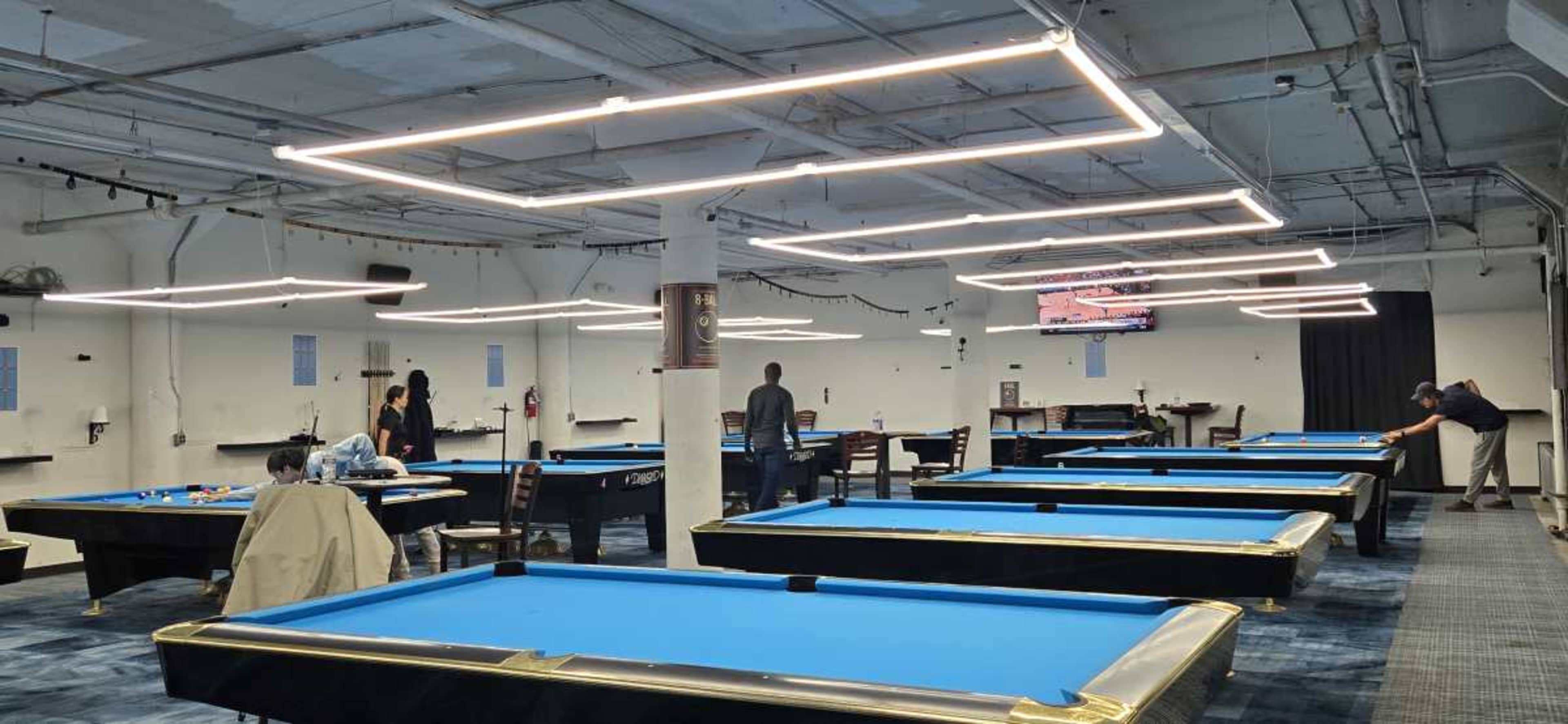 The image shows a spacious pool hall with multiple blue billiard tables and overhead lighting, where several people are playing or observing.