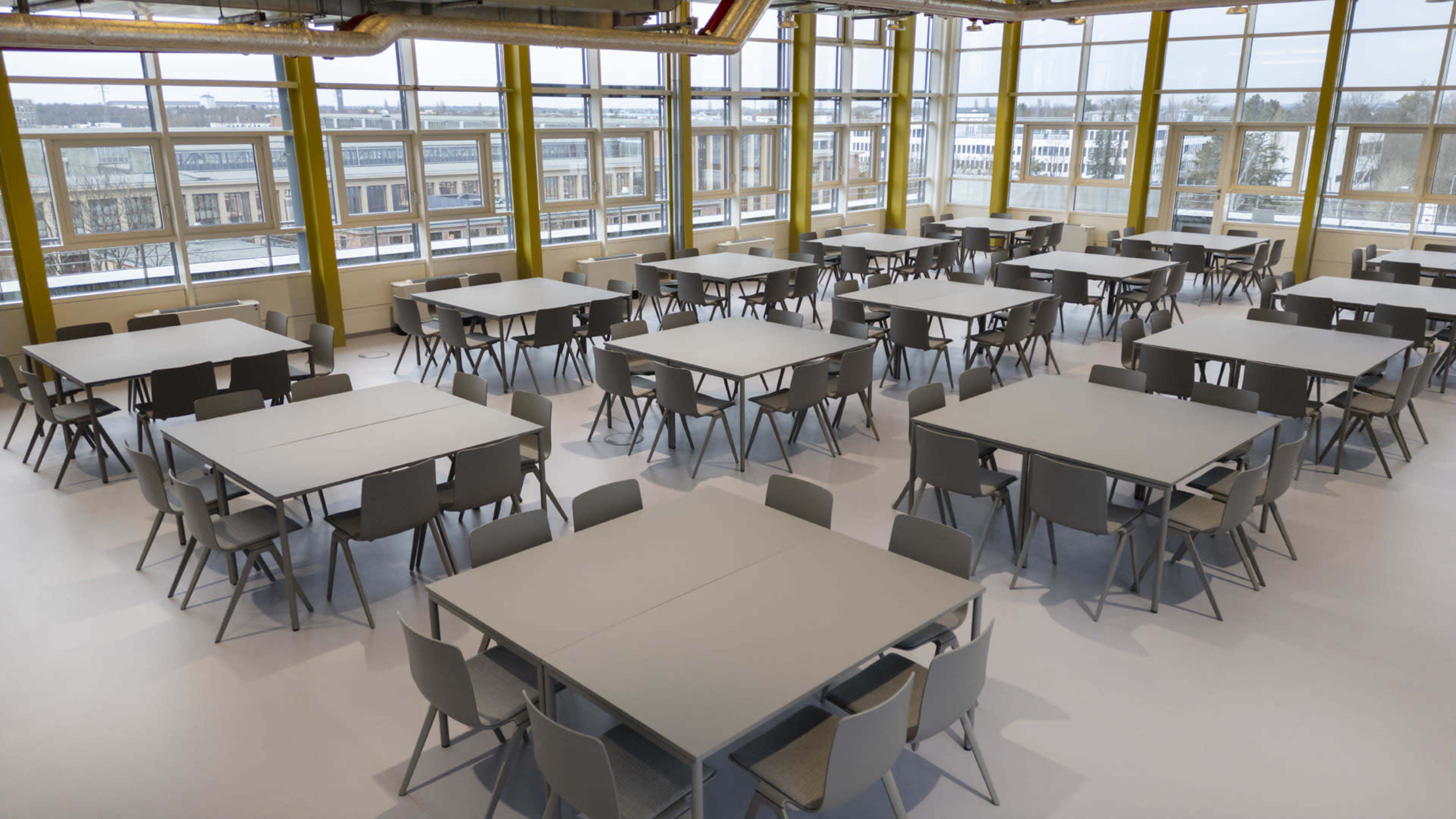 The image shows a spacious room with multiple gray tables and chairs arranged in an orderly fashion, illuminated by large windows that allow natural light to enter.