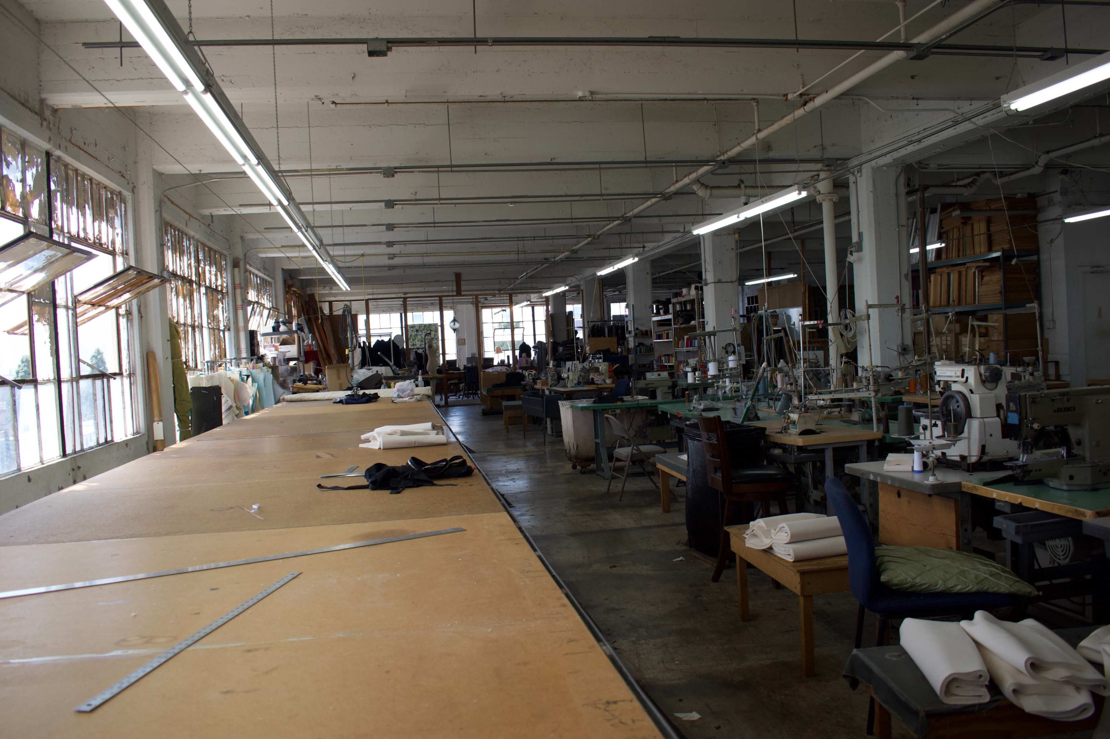 A spacious sewing workshop features large tables, various sewing machines, and ample natural light from the windows.