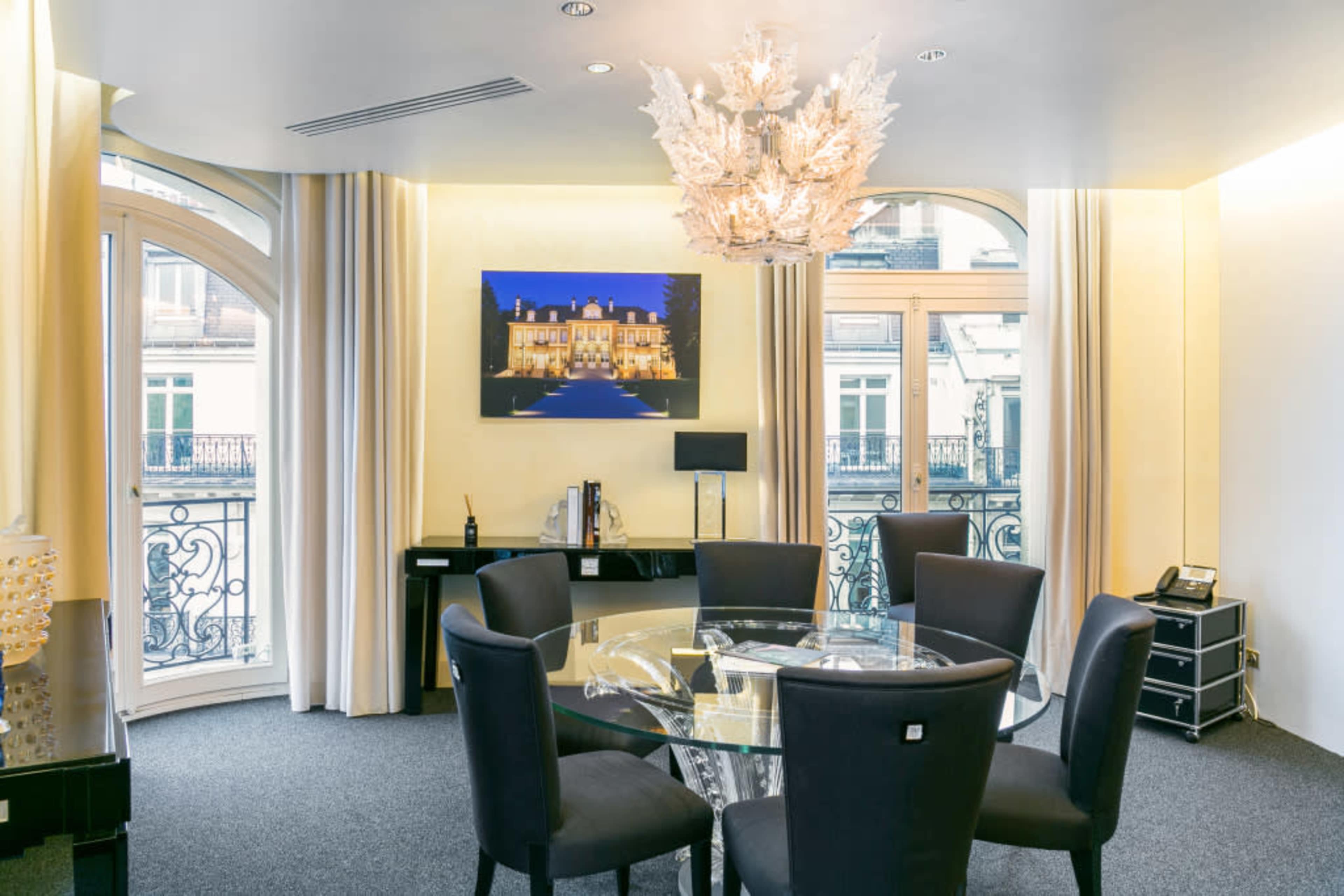 A modern meeting room with a round glass table surrounded by black chairs, illuminated by a decorative chandelier, and featuring large windows with elegant drapes.