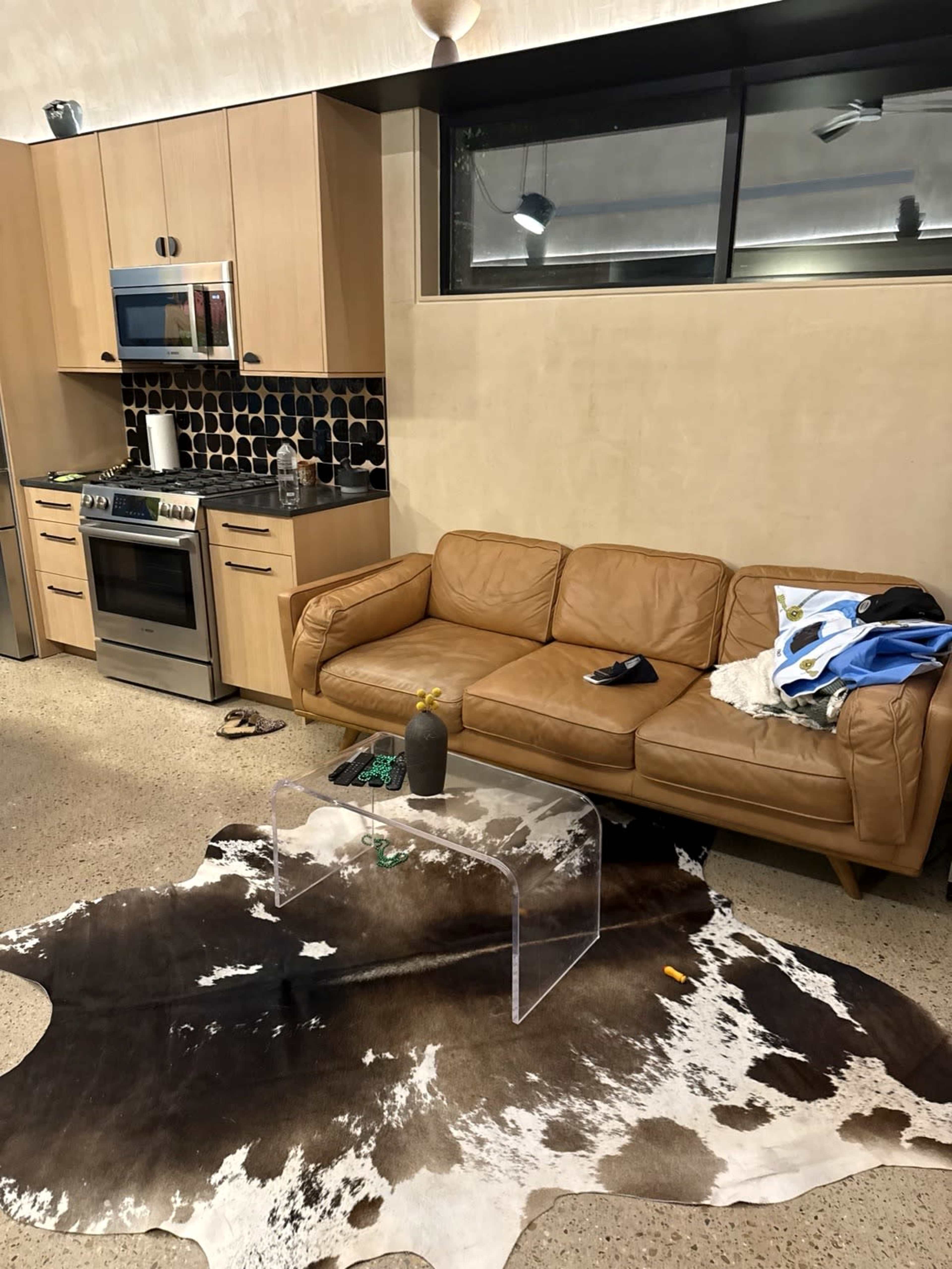 The image shows a modern living space with a brown leather couch, a clear acrylic coffee table on a cowhide rug, and a kitchen area featuring stainless steel appliances and dark tiled backsplash.