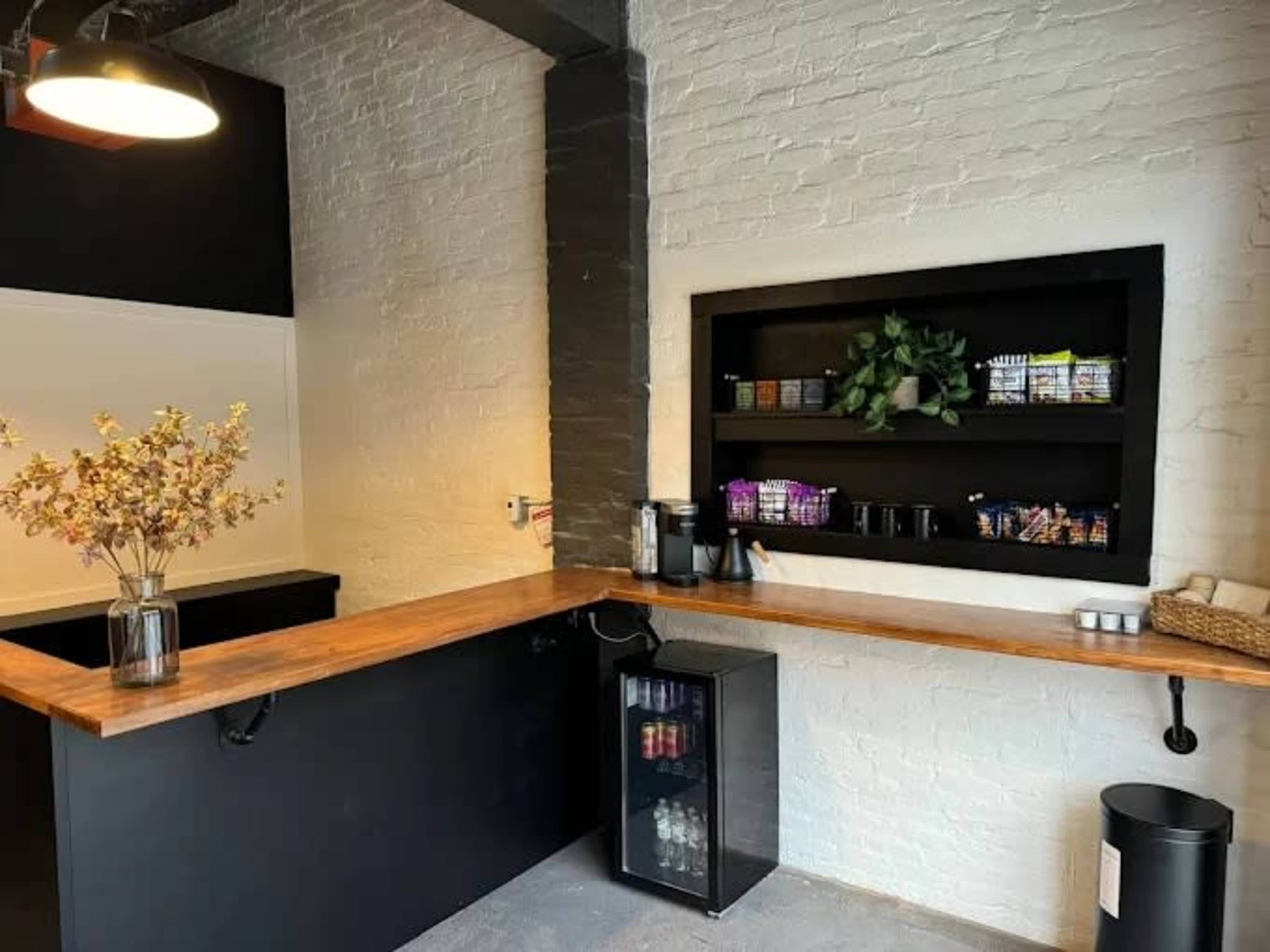 The image shows a minimalist café area with a wooden countertop, a small black refrigerator, and a shelf featuring various snack items and a potted plant.