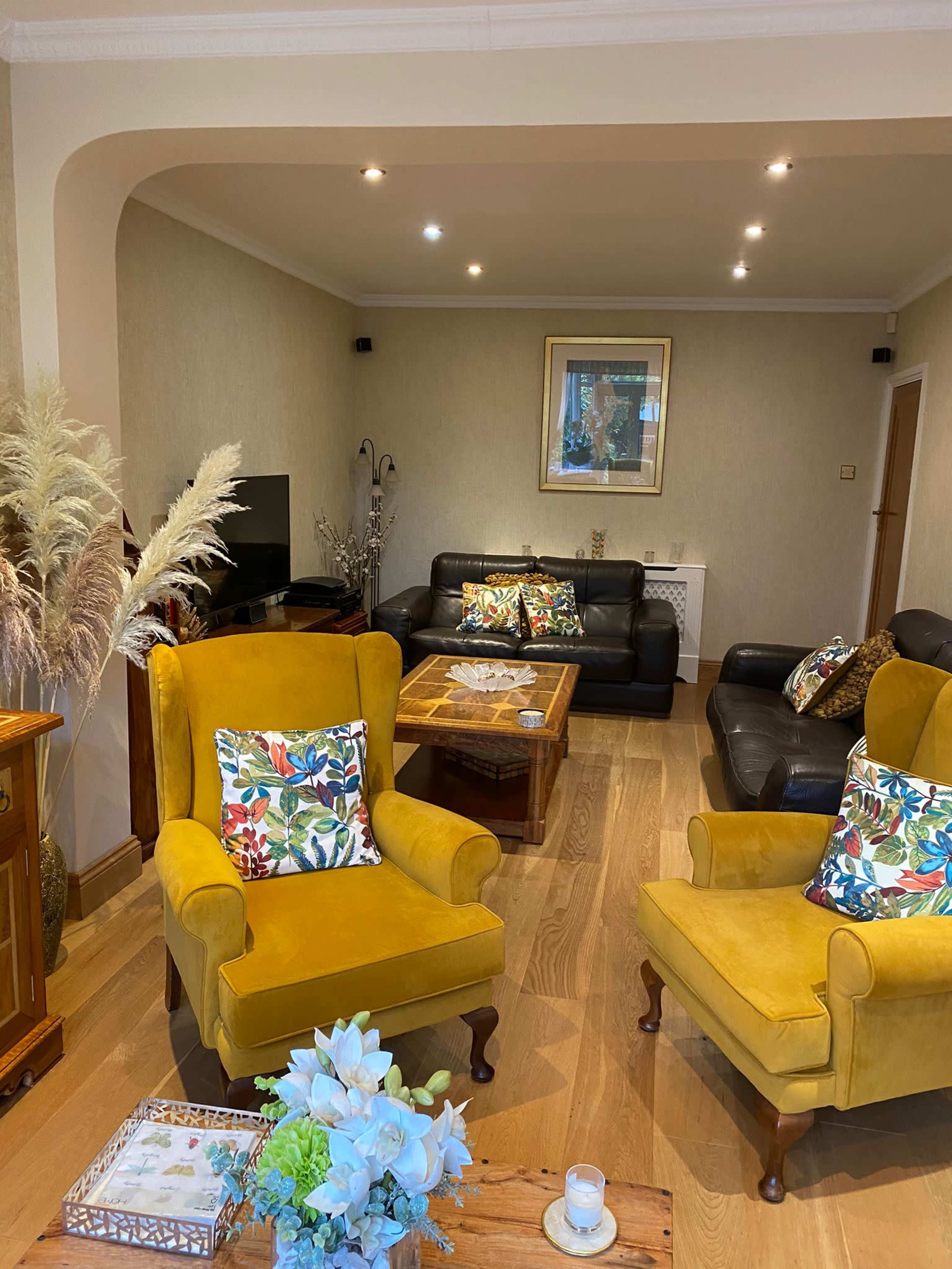 A living room features two yellow armchairs, a glass coffee table, and a sofa set, all arranged around a television and decorative plants.