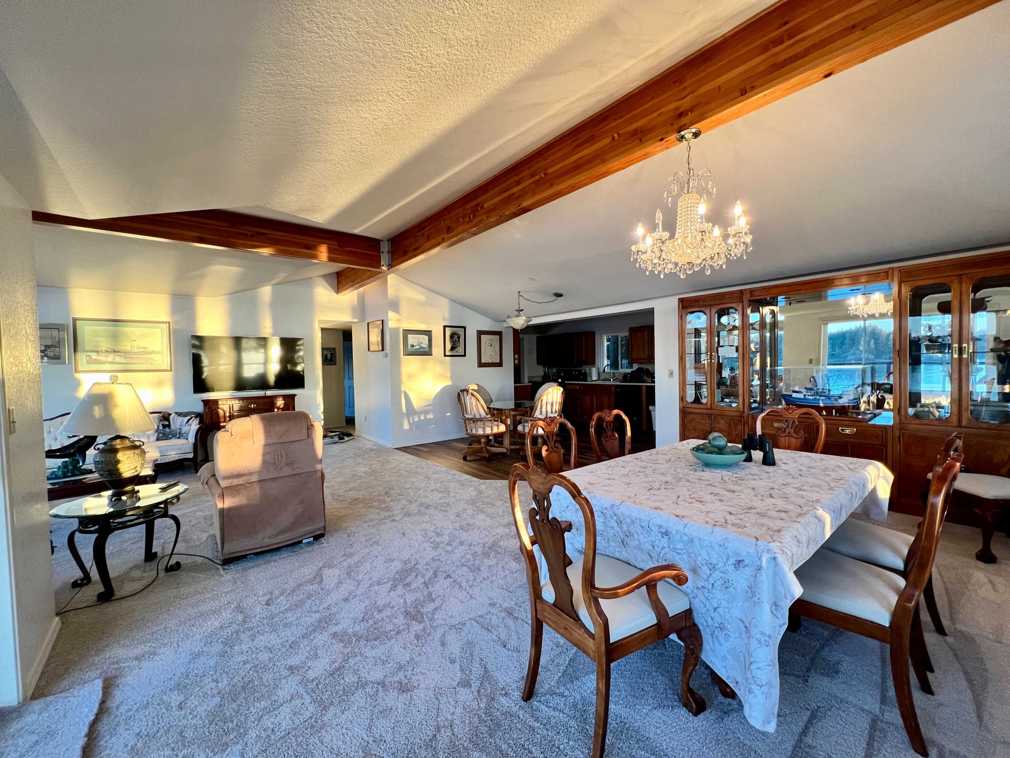 The image shows a spacious living and dining area with a chandelier overhead, wooden beams, and a set dining table near a display cabinet.
