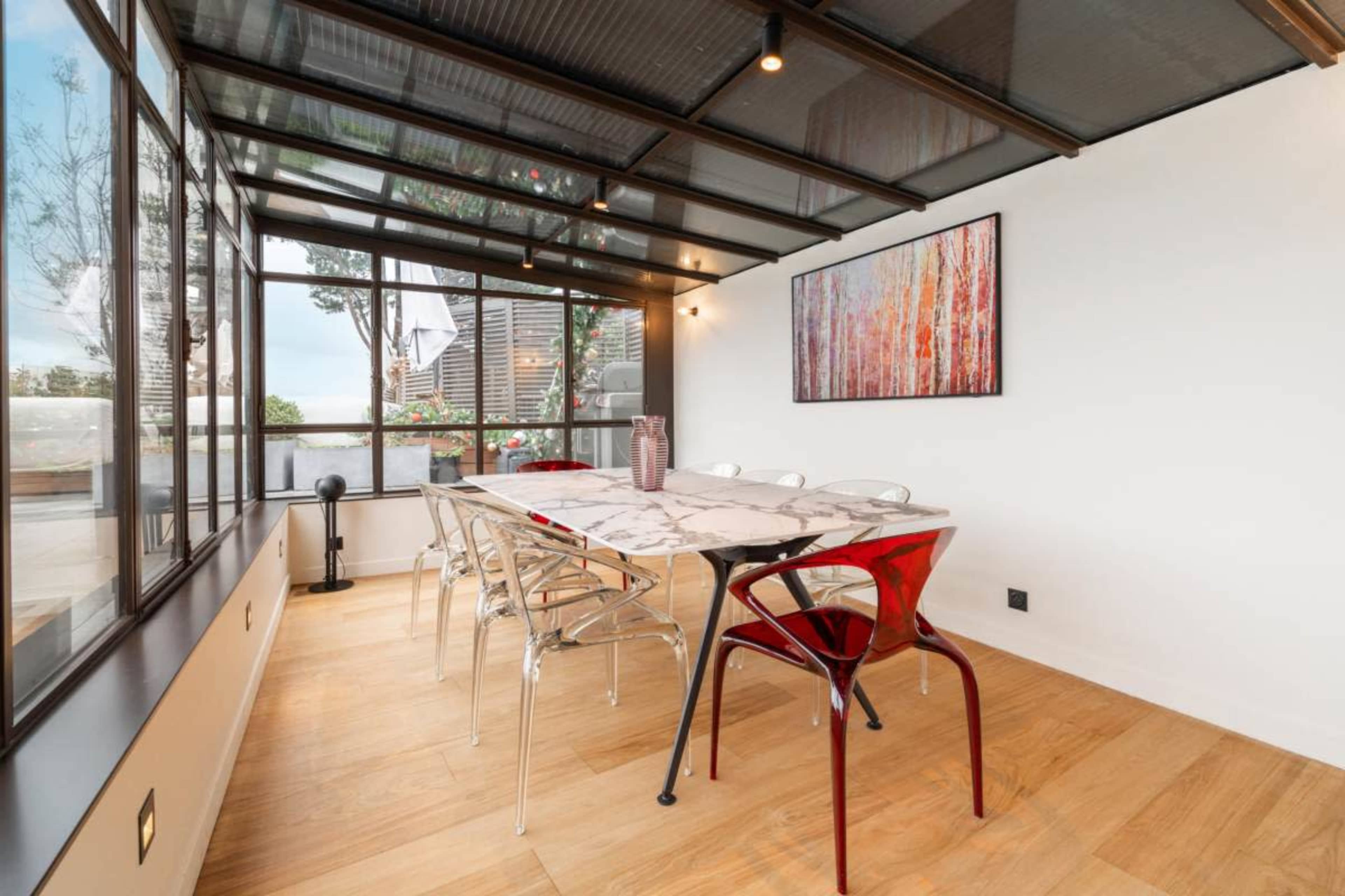 The image shows a modern dining area with a glass roof, featuring a long marble table surrounded by transparent and red chairs, and large windows overlooking an outdoor space.