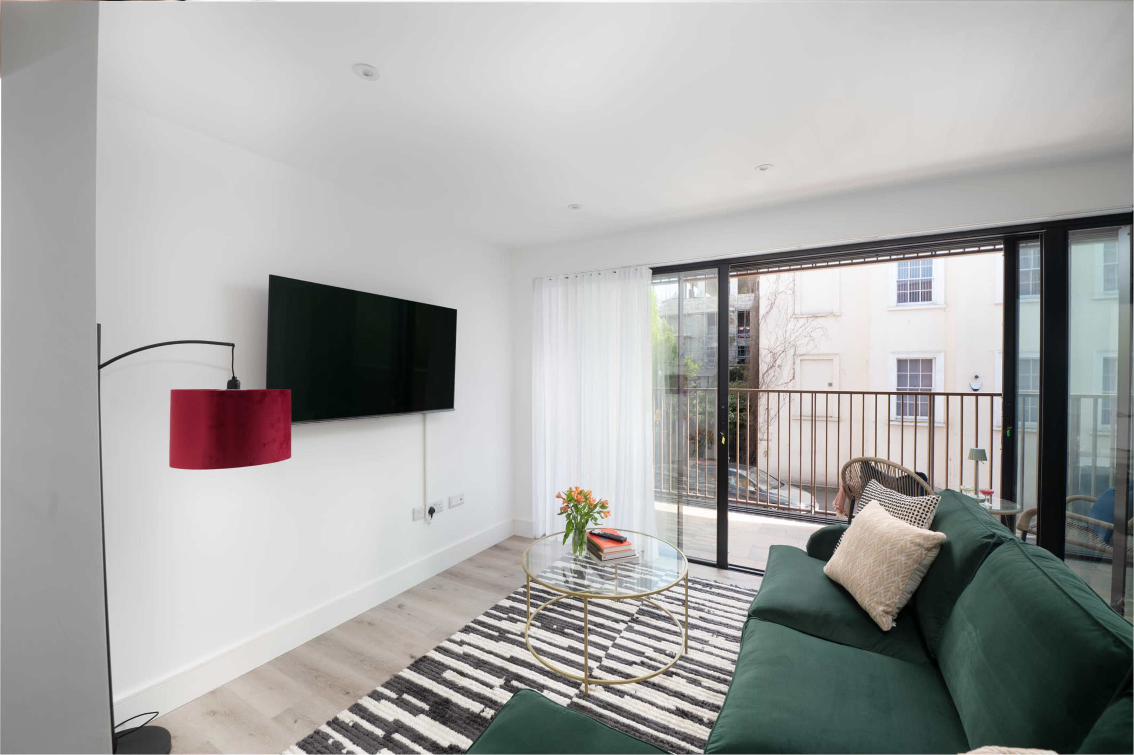 A modern living room features a green sofa, a round glass table, a large wall-mounted television, and sliding glass doors opening to a balcony.