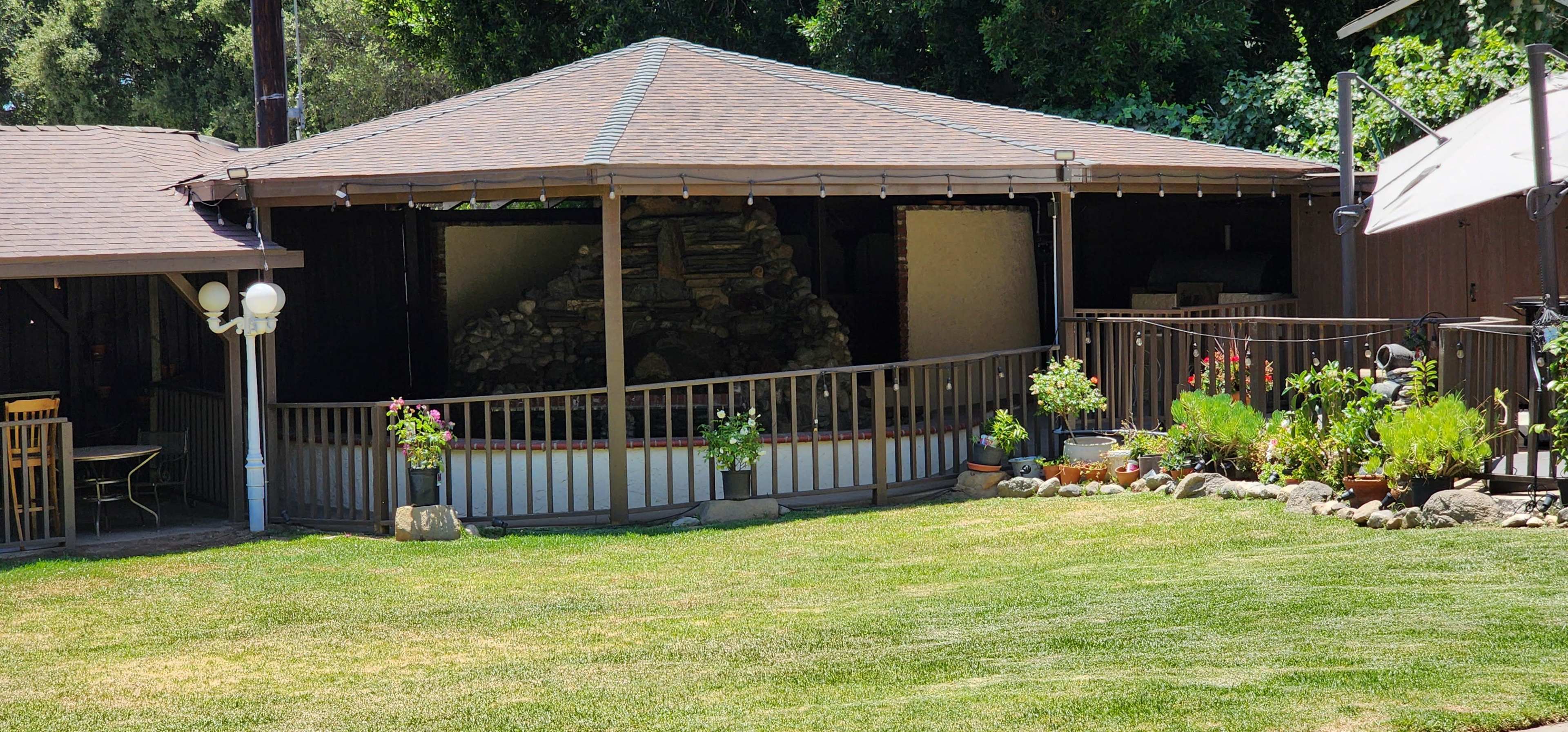 A circular wooden pavilion with a peaked roof is surrounded by a grassy area and contains potted plants along the railing.