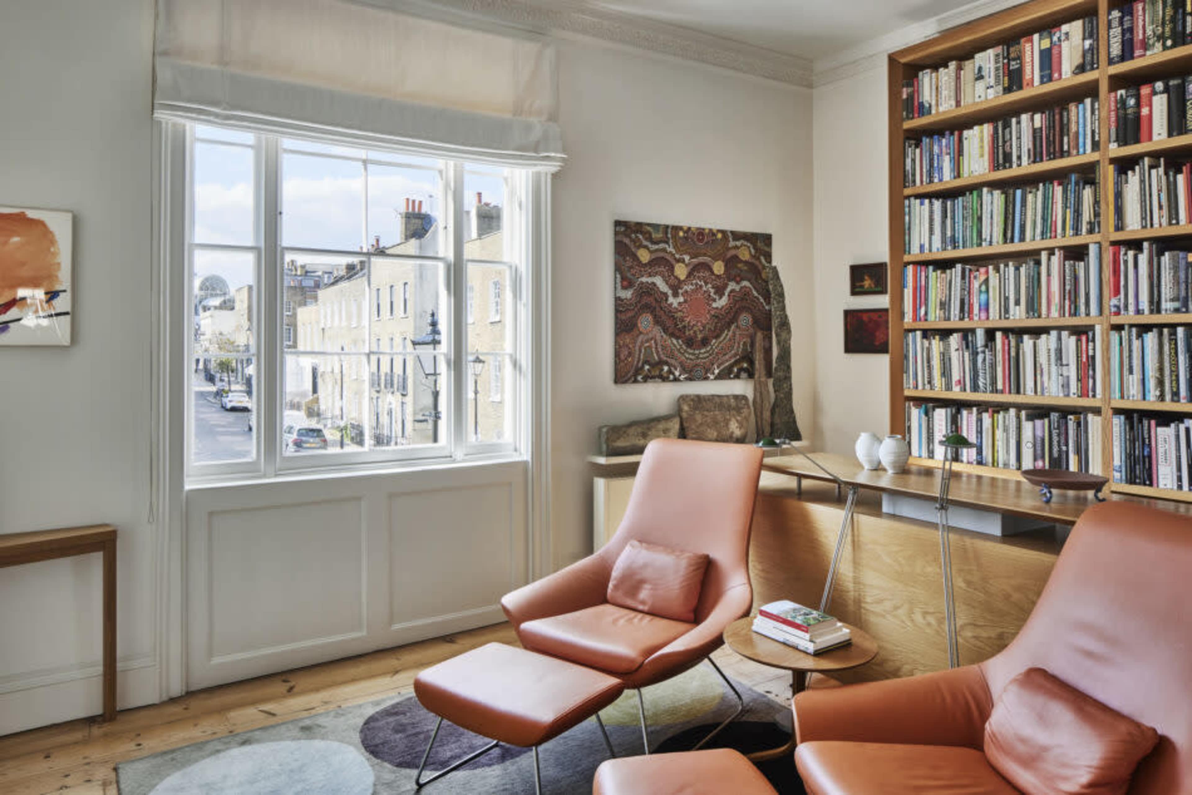 A cozy interior features two orange chairs, a wooden desk, and a large bookshelf beside a window overlooking a street.