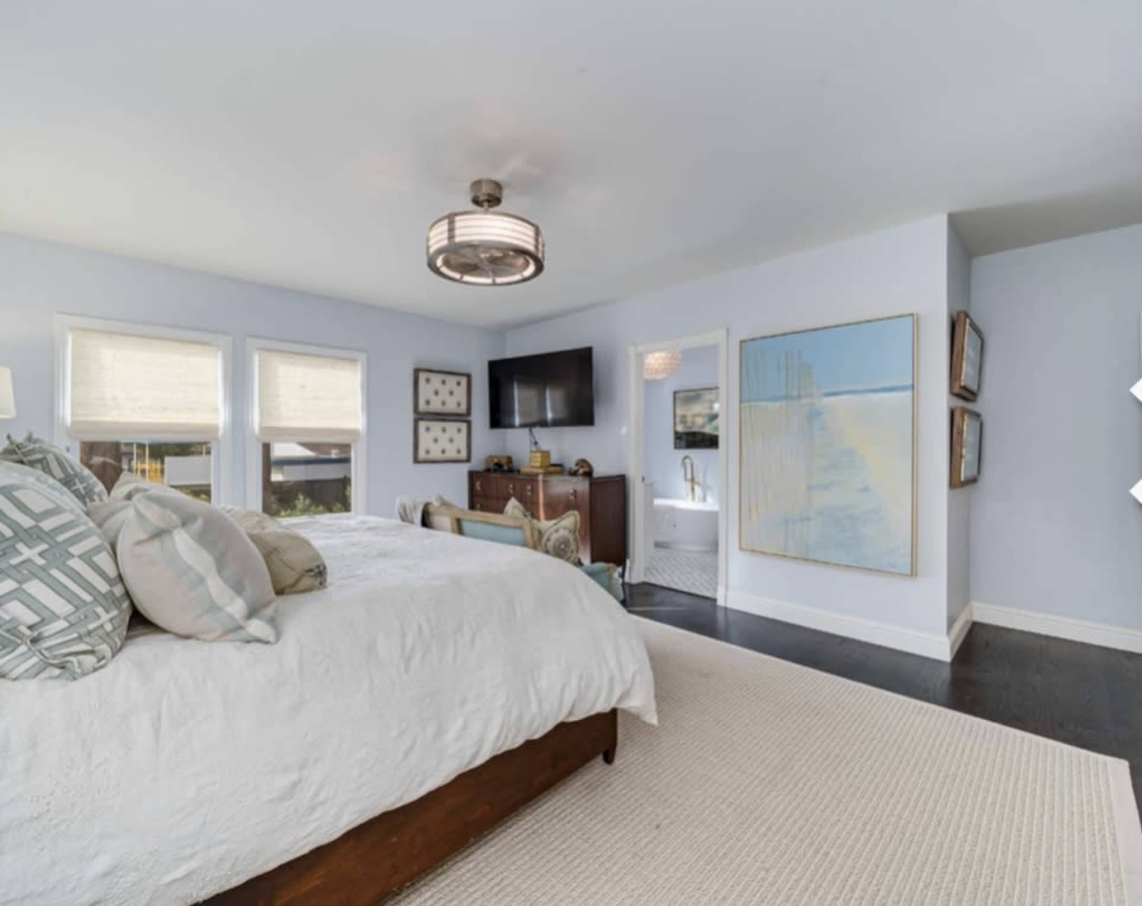 A spacious bedroom features a large bed, wall-mounted television, and abstract art, with natural light filtering through shades.
