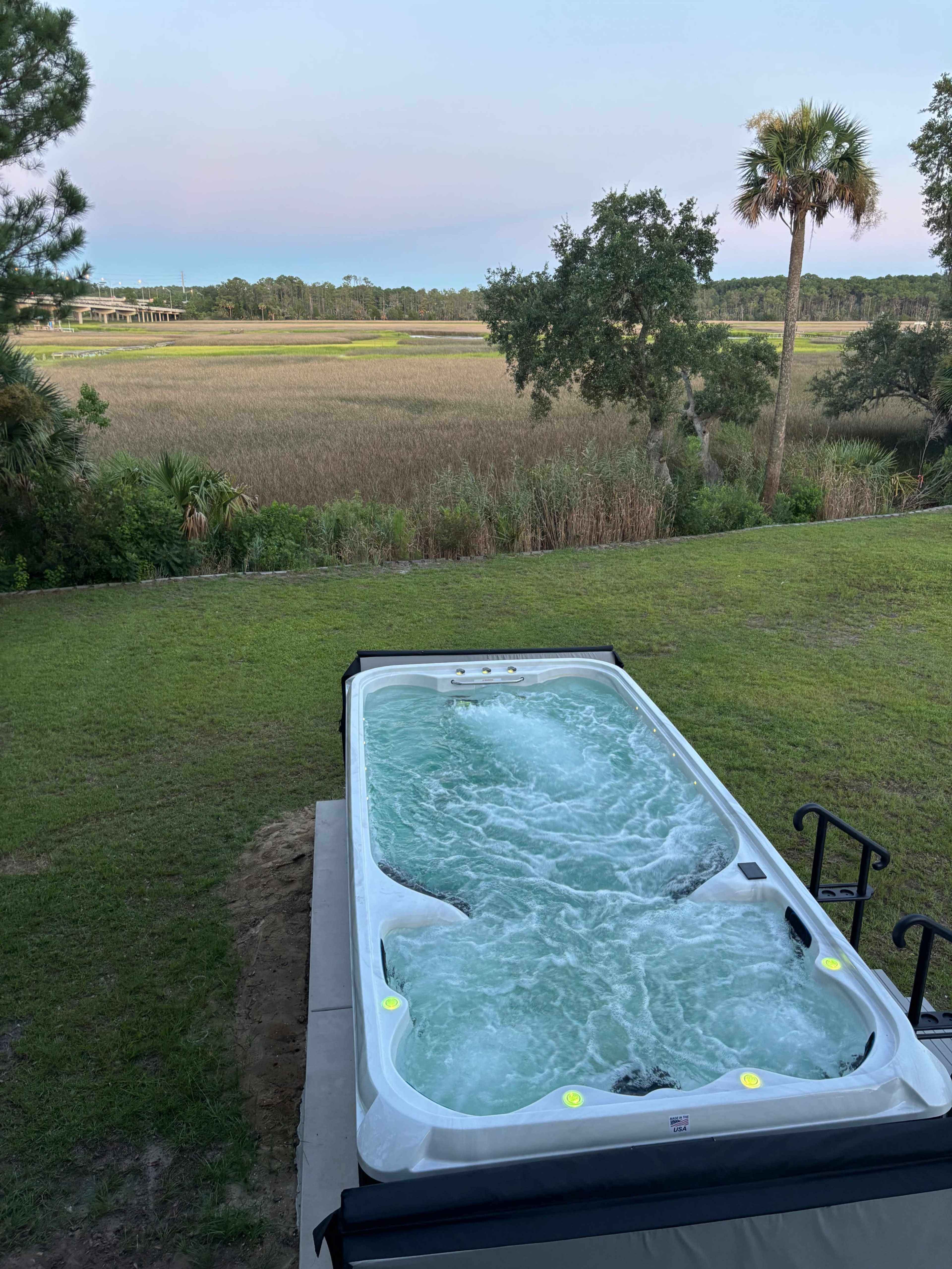 A hot tub filled with bubbling water sits on a grassy area overlooking a marshland landscape.