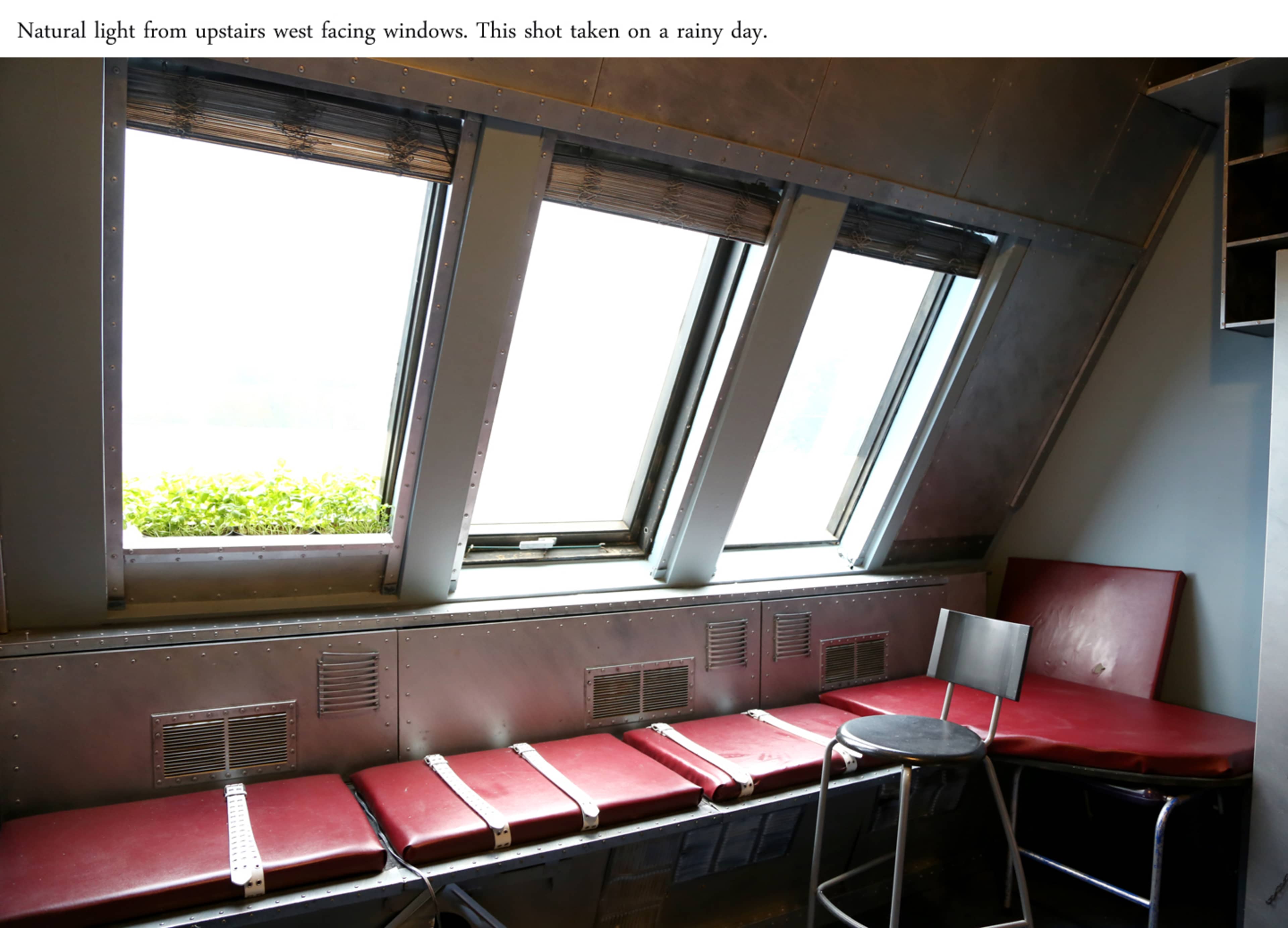 The image shows a seating area with red chairs and a barstool next to large, west-facing windows that let in natural light on a rainy day.