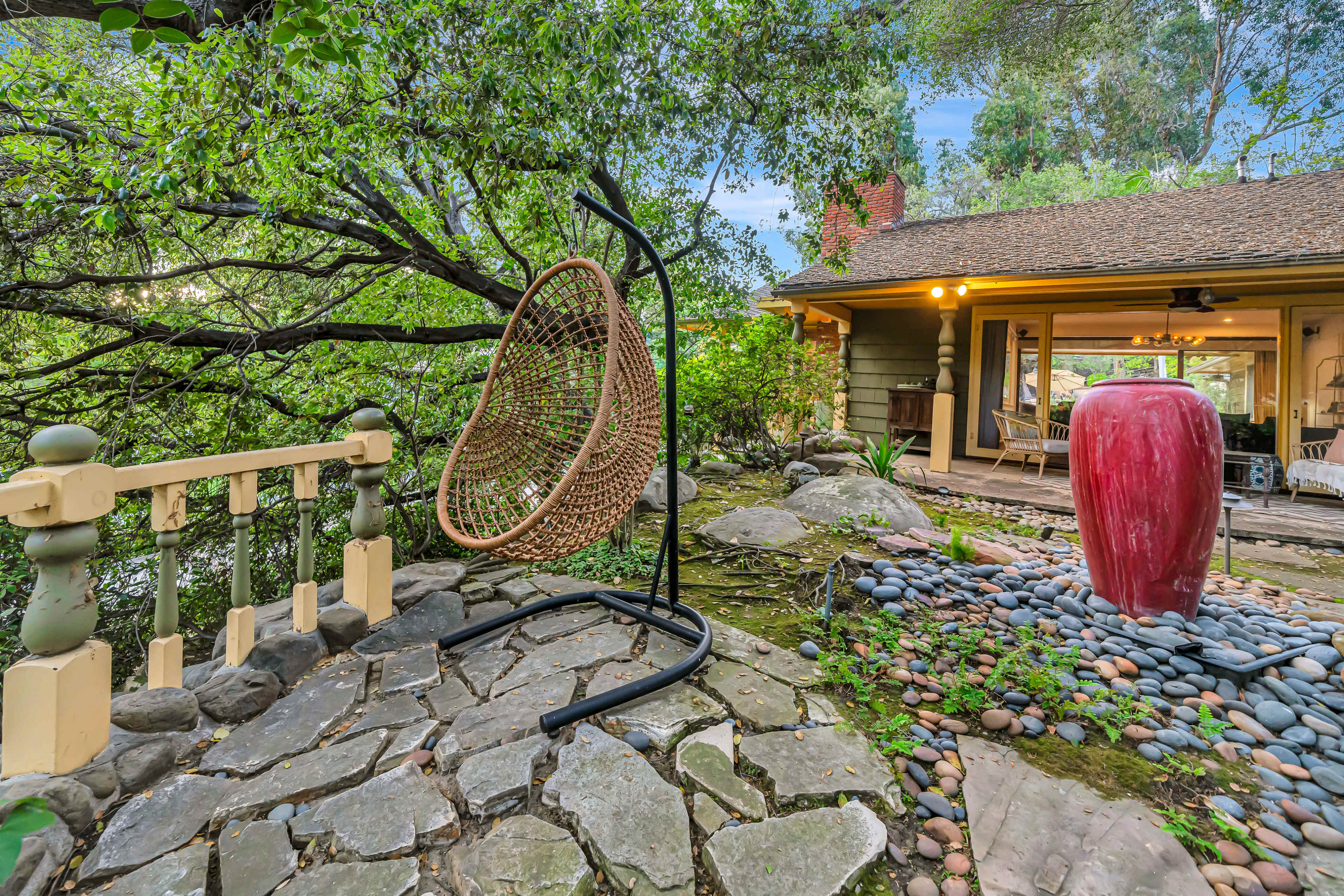 A hanging wicker chair is situated beside a stone path and a red sculpture, with a cabin visible in the background.