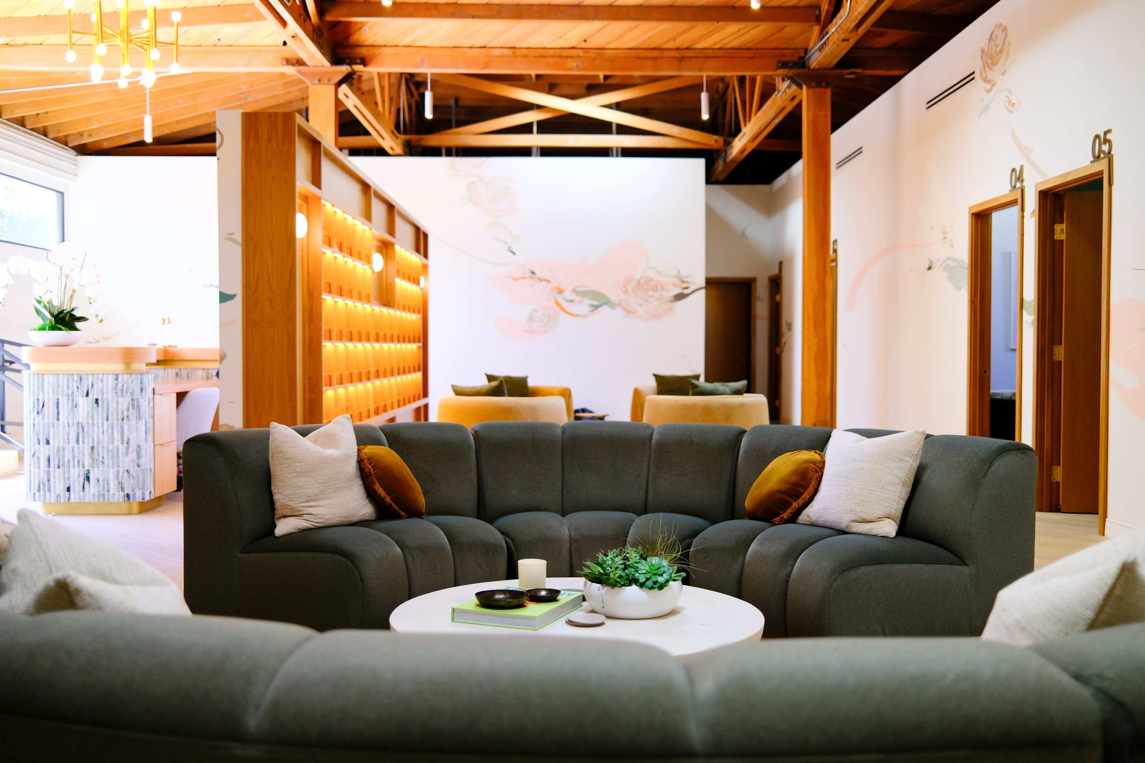 The image shows a modern, open lounge area featuring a curved gray sofa arranged around a circular coffee table, with wooden beams visible on the ceiling and decorative elements on the walls.