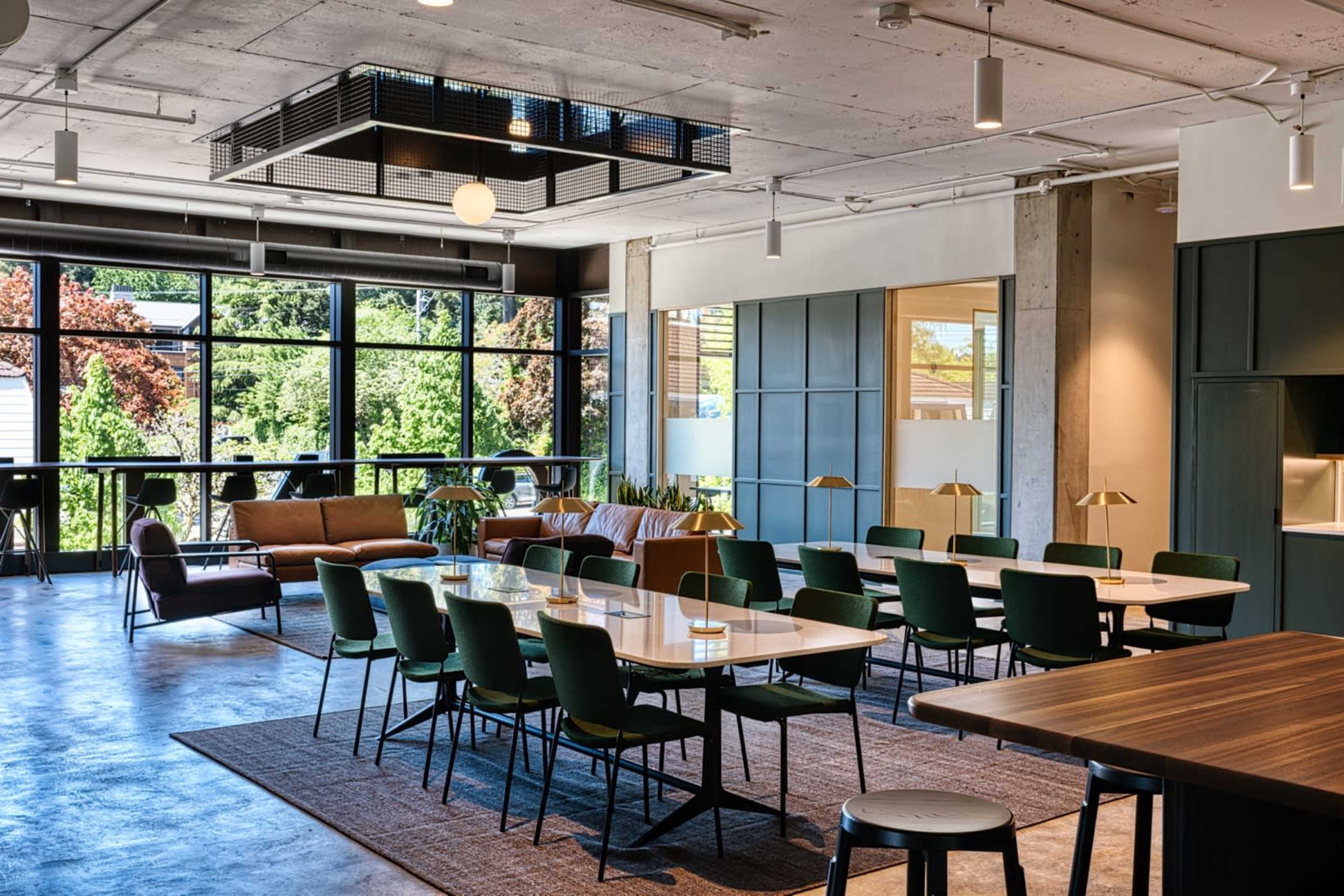 The image depicts a modern open-plan workspace with a seating area and a dining table surrounded by green chairs, large windows allowing natural light, and minimalistic decor.