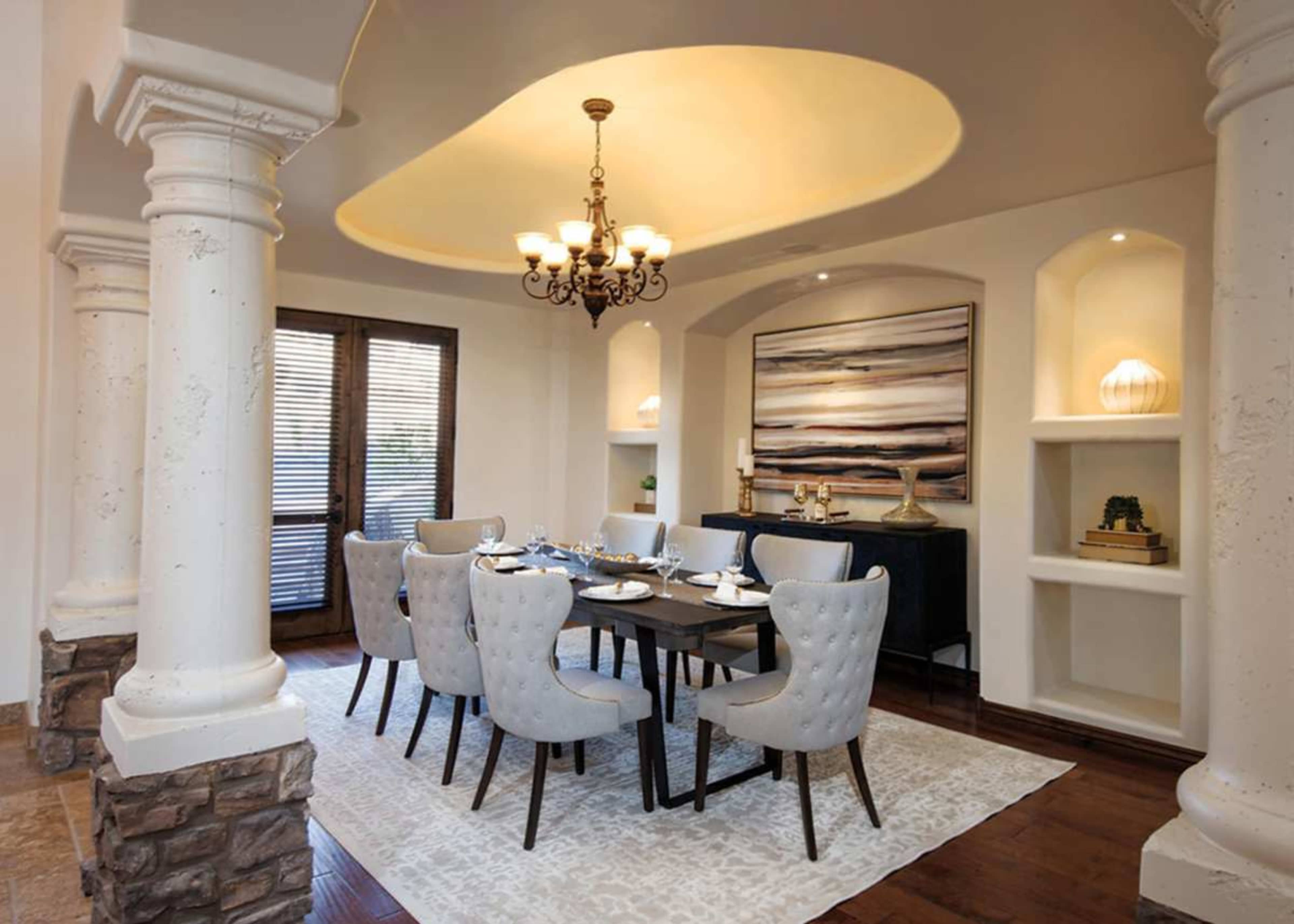 The image features a spacious dining room with a large wooden table surrounded by upholstered chairs, accented by a chandelier and decorative wall art.