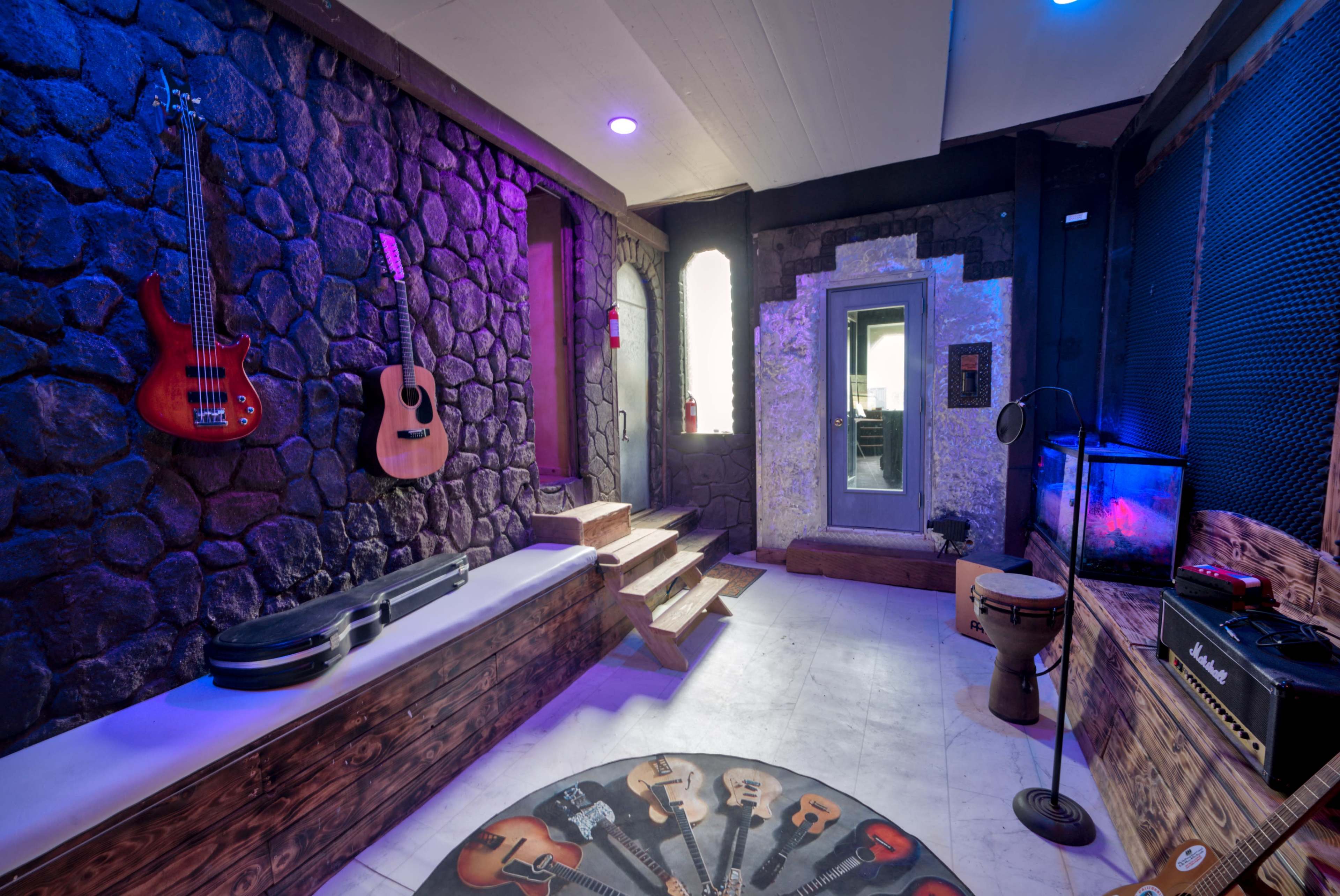 The image shows a music rehearsal space featuring stone walls, a staircase, various guitars displayed, and a soundproof door.