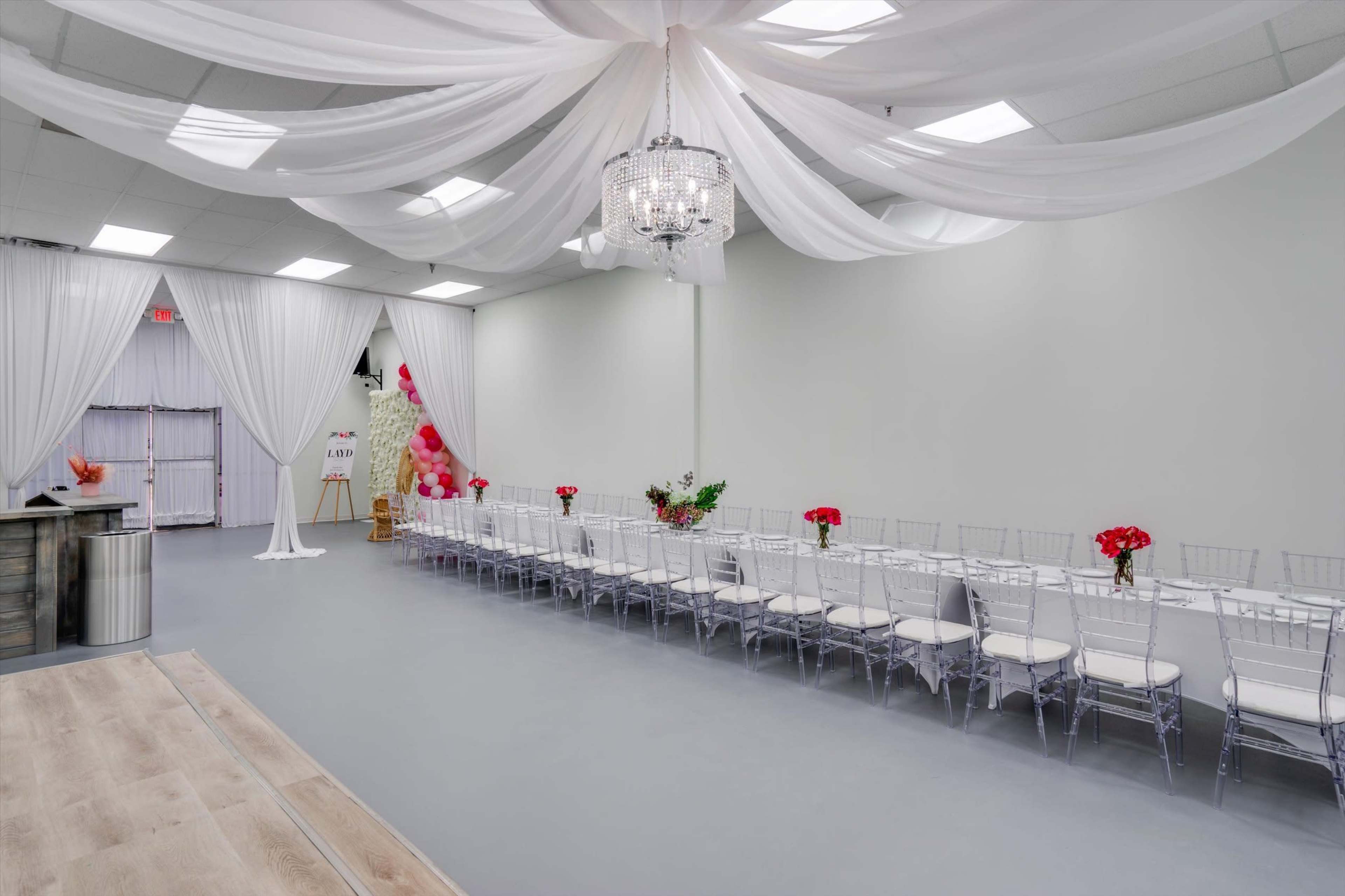 A spacious event room decorated with white drapery and a chandelier, featuring a long table set with chairs and floral arrangements.