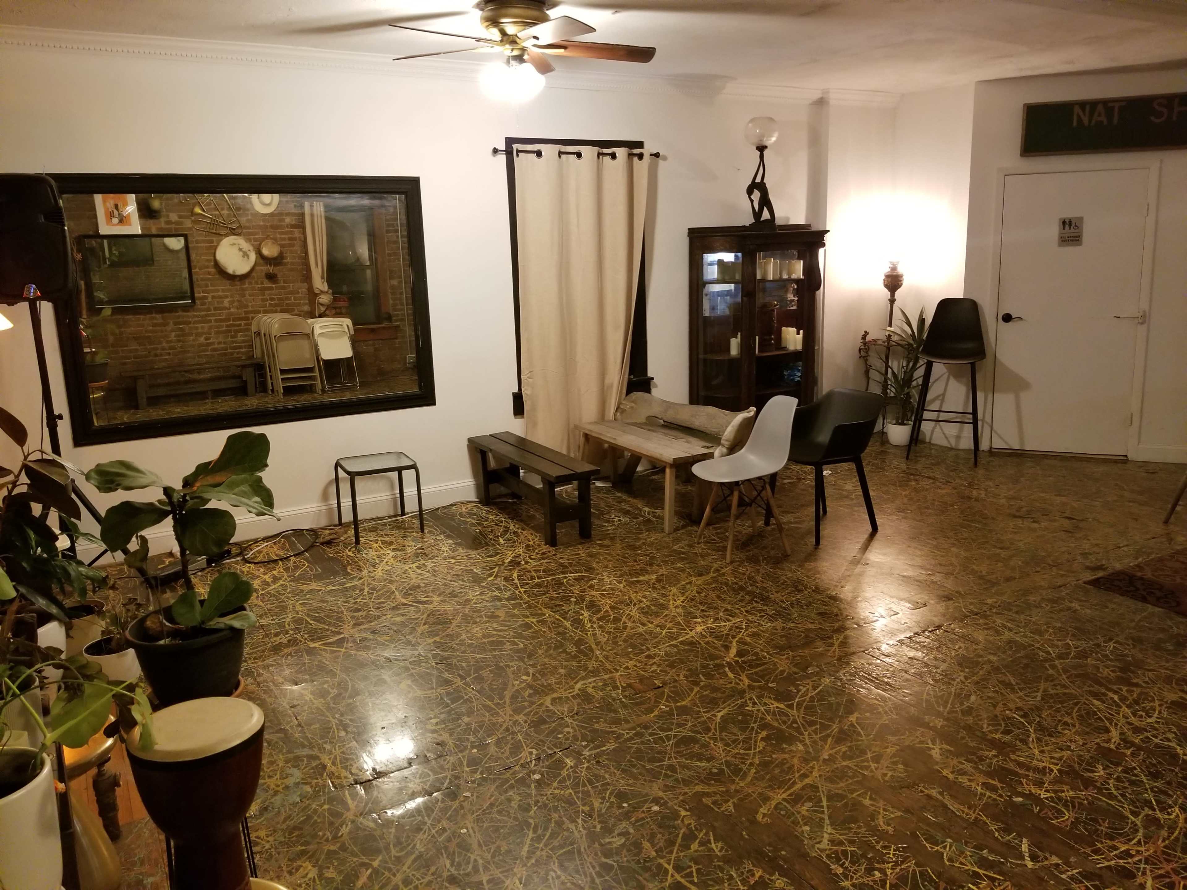 The interior of a room features wooden flooring, a large mirror, various chairs, a curtain, and a display cabinet against a brick wall.