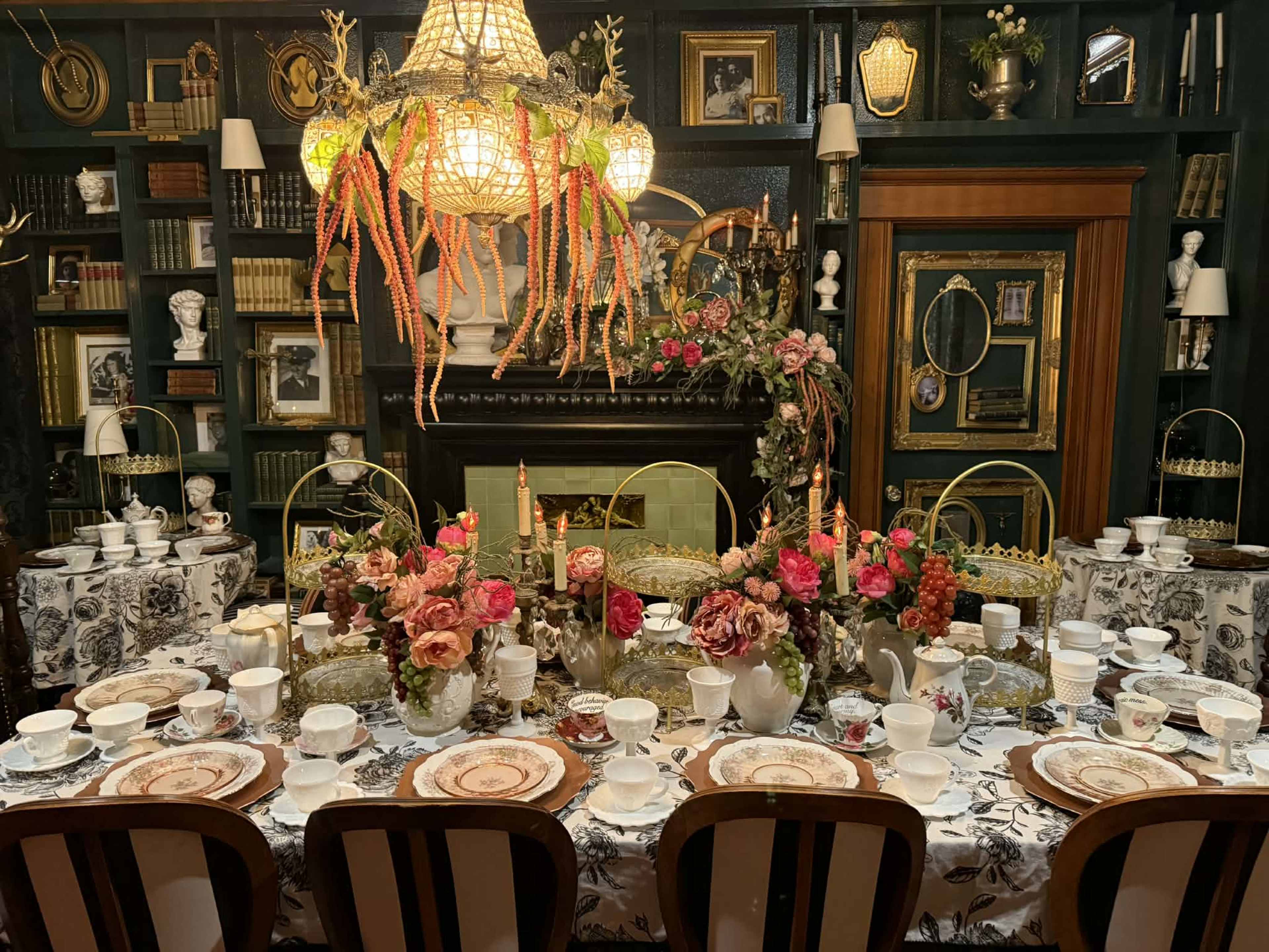 A beautifully set dining table with fine china, floral arrangements, and vintage decor is displayed in a room filled with books and framed portraits.