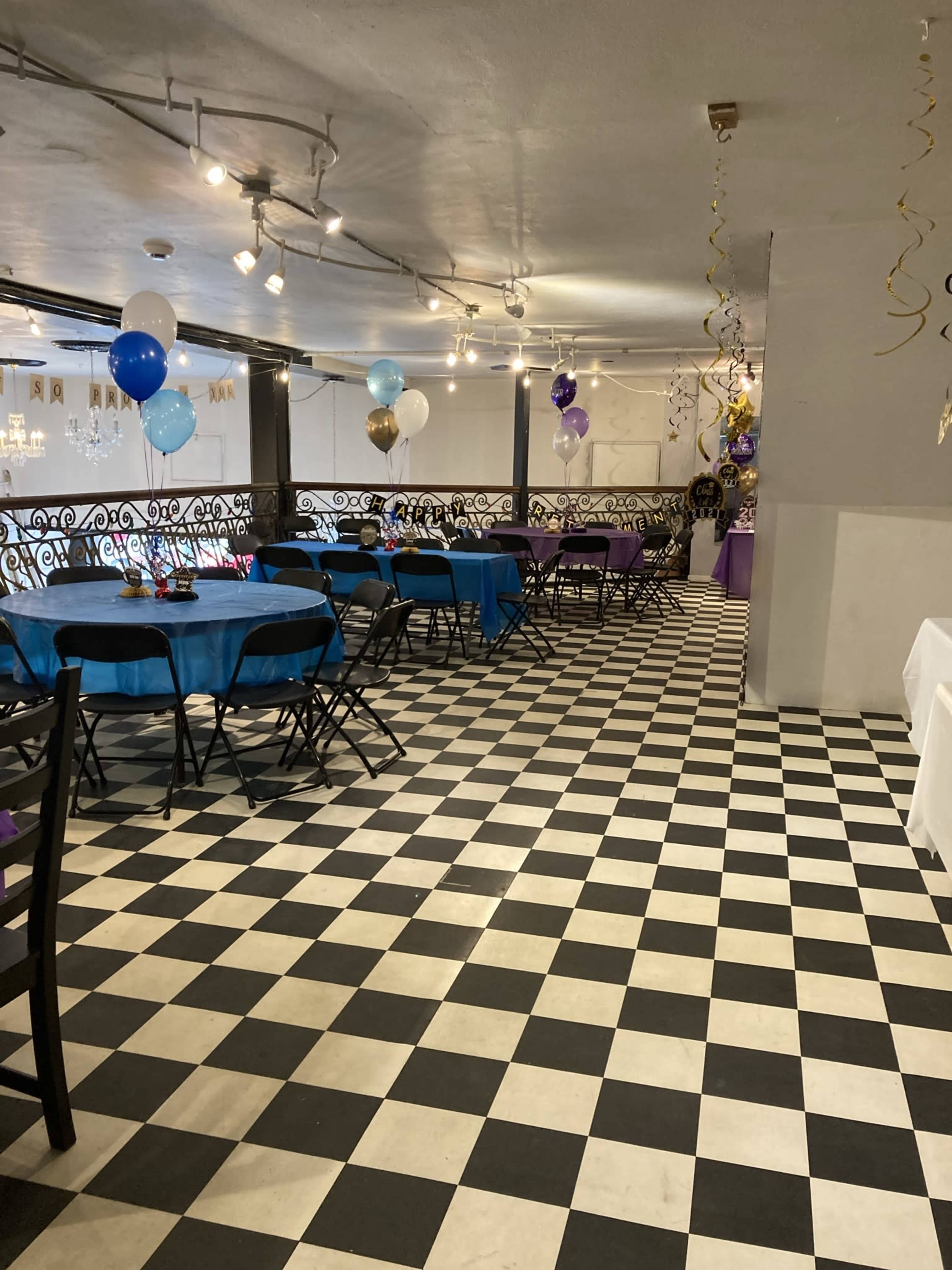 The image shows a large indoor space set up for a celebration, featuring black and white checkered flooring, blue and purple table covers, and colorful balloons.