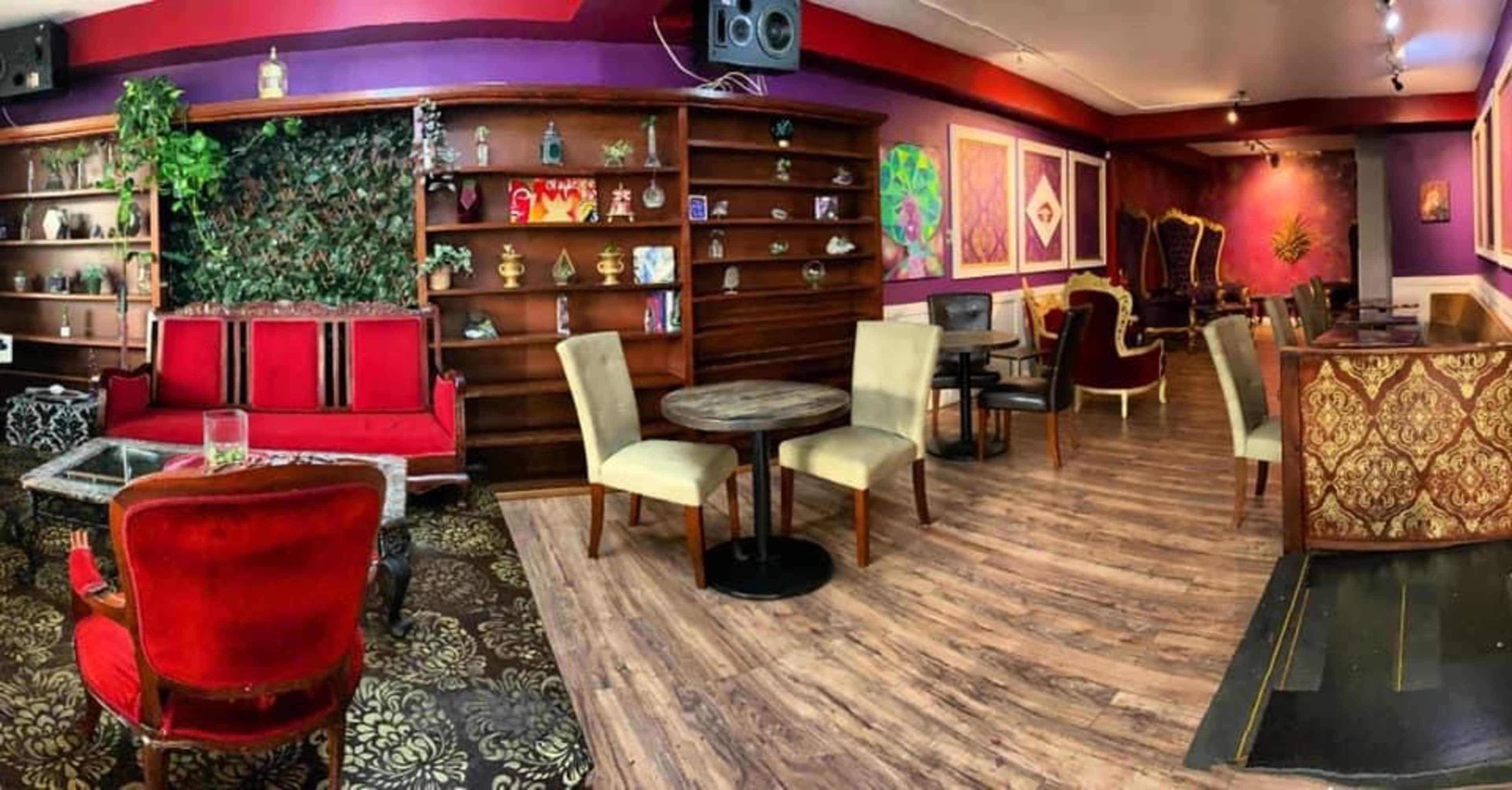 The image shows a cozy café interior with red and gold furniture, wooden shelves lined with decorative items, and a mix of seating arrangements including tables and chairs.