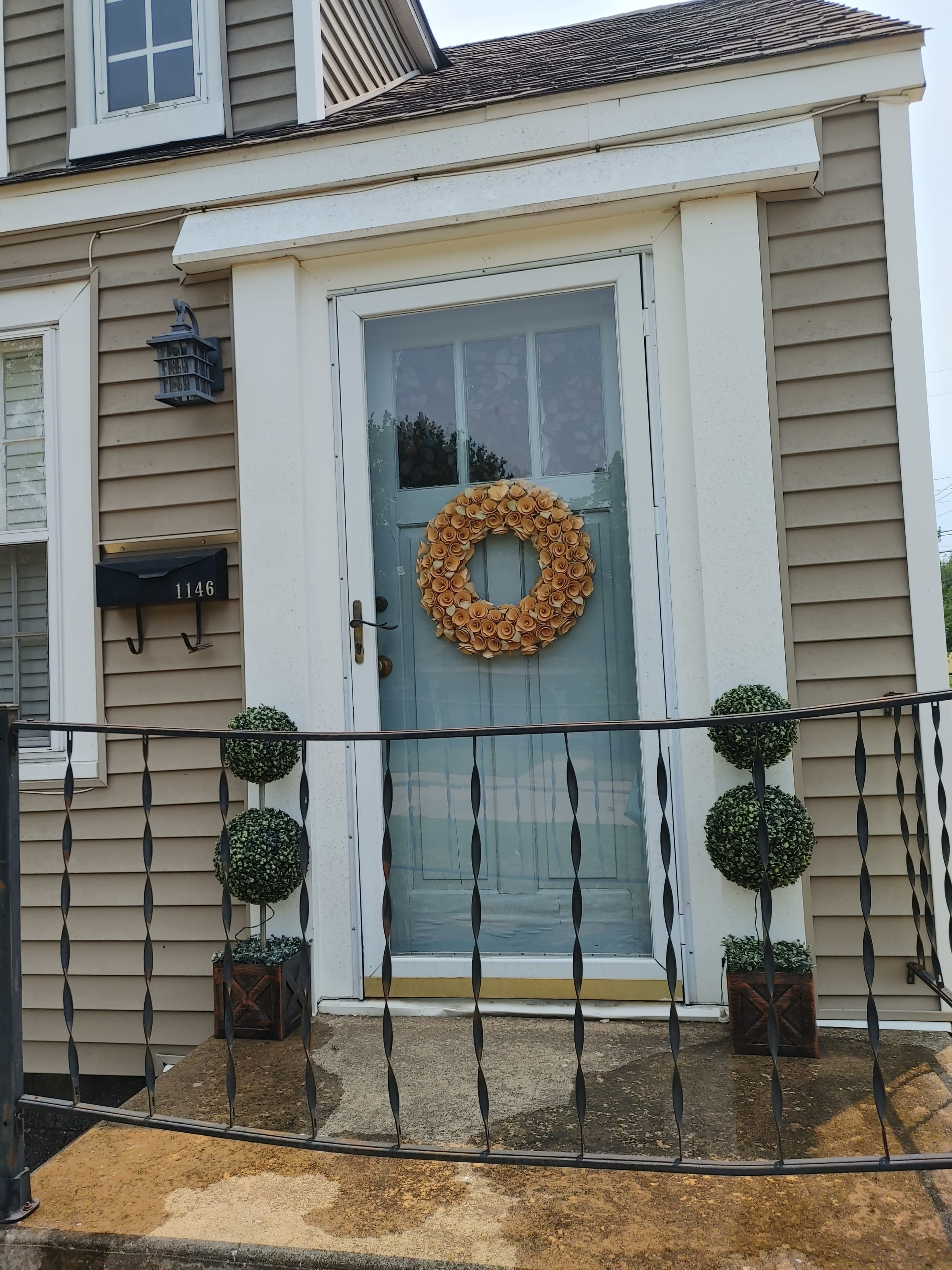 A front door with a decorative wreath is framed by two potted topiary plants on a small porch.