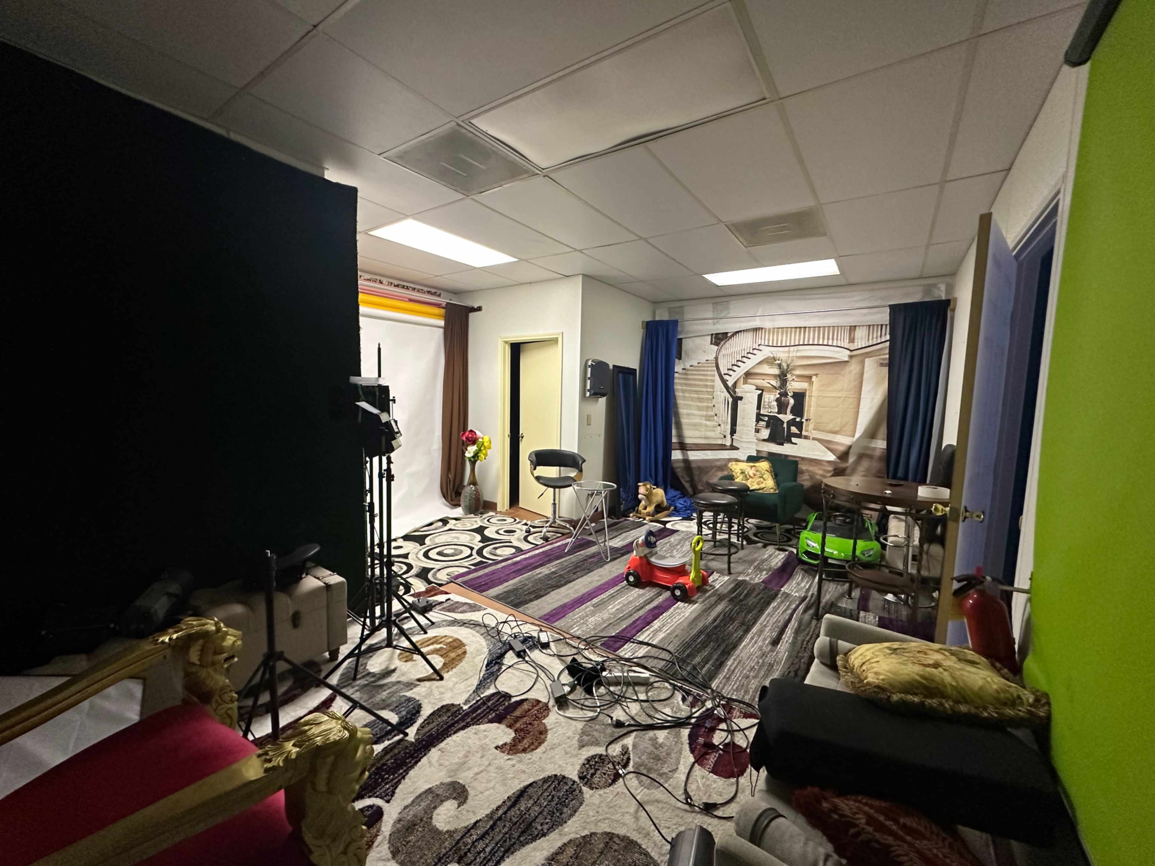 The image shows a studio space with a colorful rug, a backdrop of a staircase, chairs, lighting equipment, and various props arranged throughout the room.