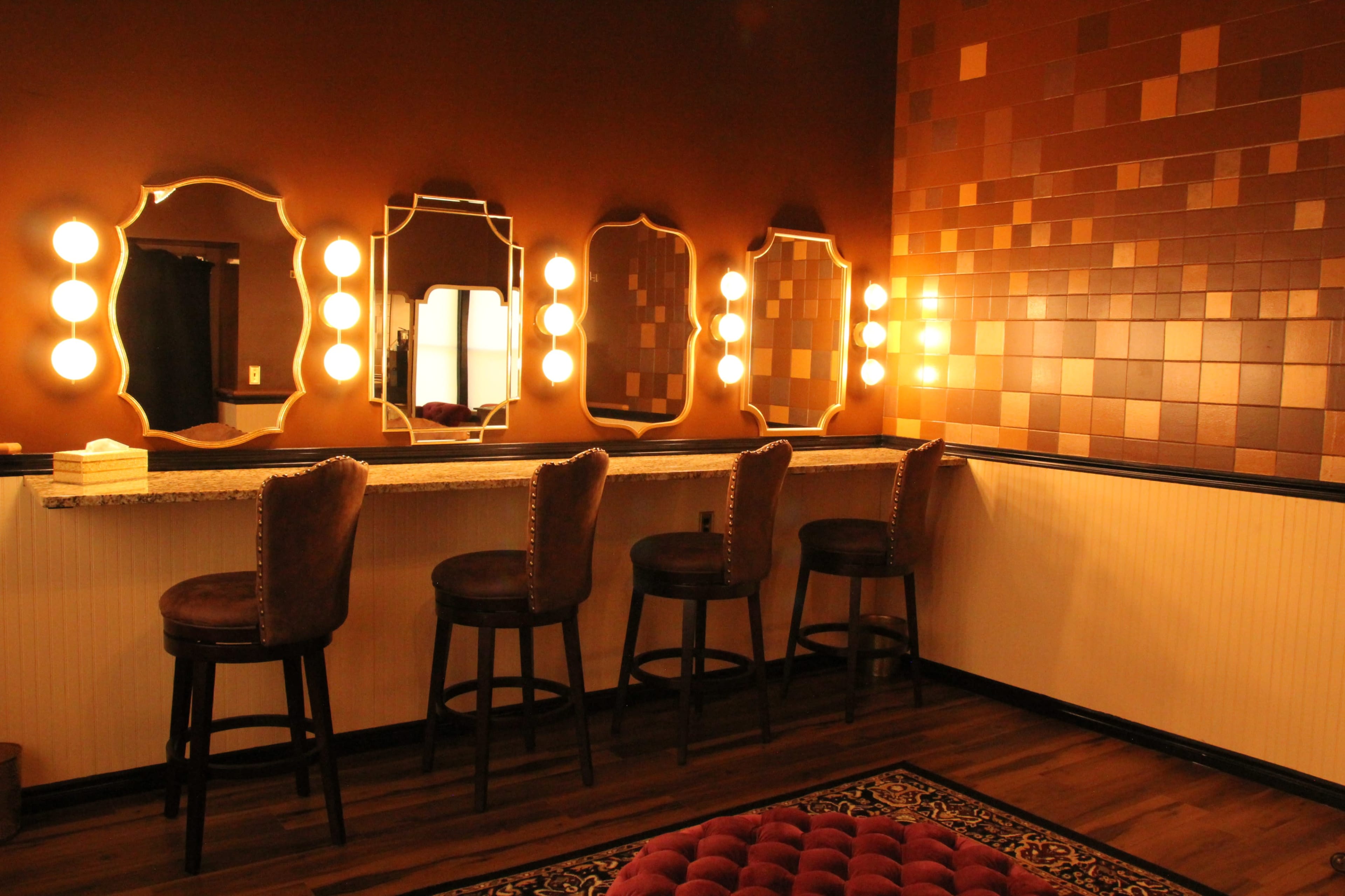 The image shows a dimly lit vanity area with a long countertop, five round vanity mirrors, and barstools set against a textured brown wall.