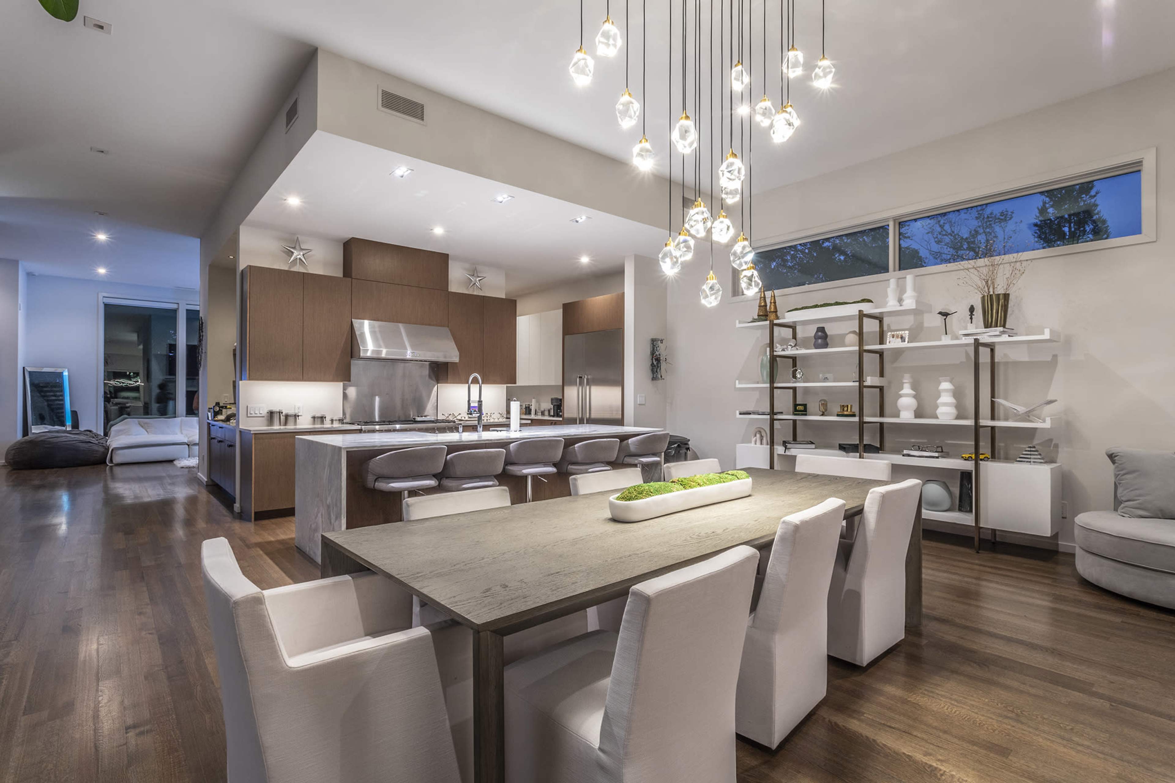 A modern dining area features a long table surrounded by upholstered chairs, with a spacious kitchen visible in the background and hanging light fixtures above.