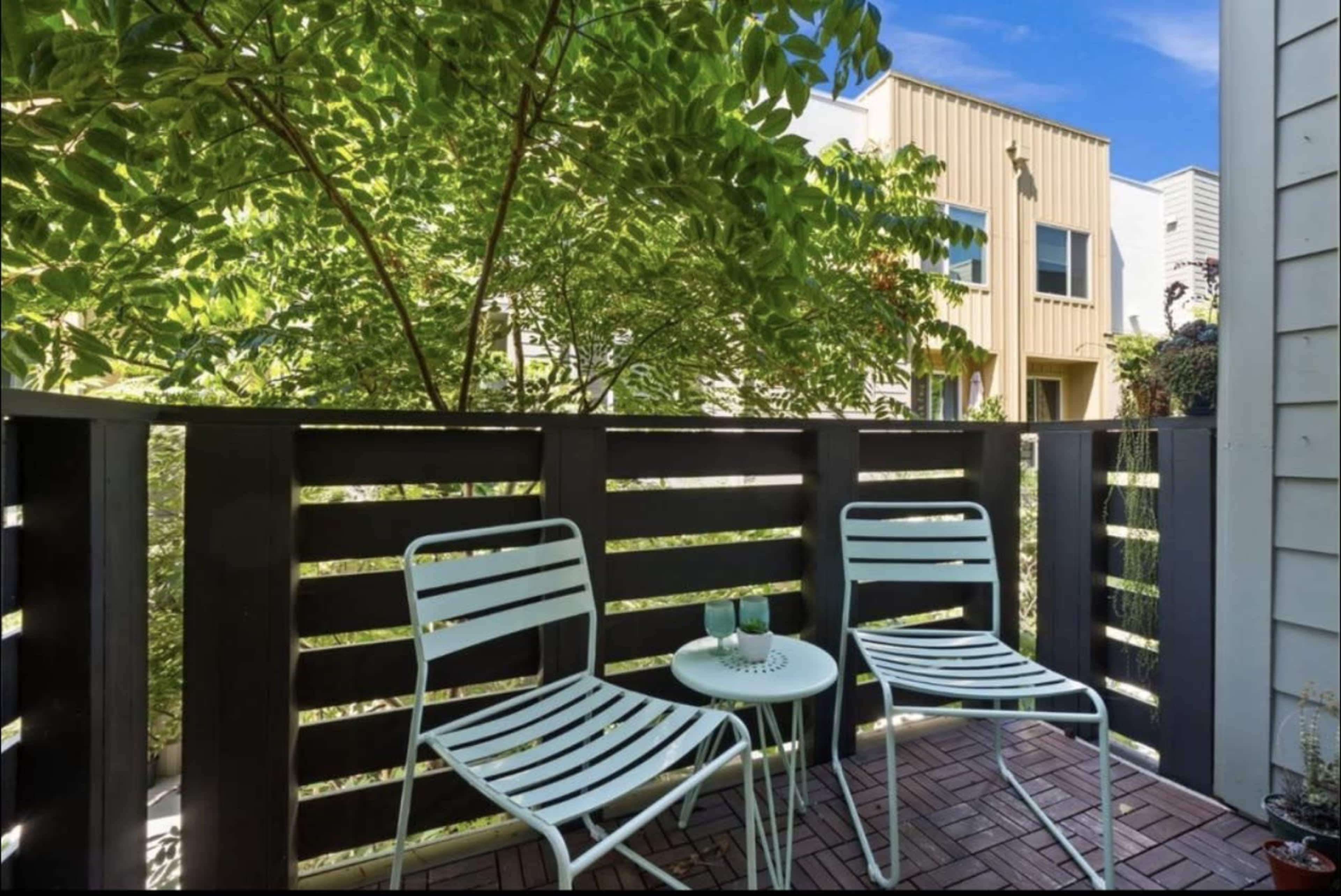 The image shows a small outdoor balcony furnished with two white metal chairs and a round table, surrounded by greenery and a view of nearby buildings.