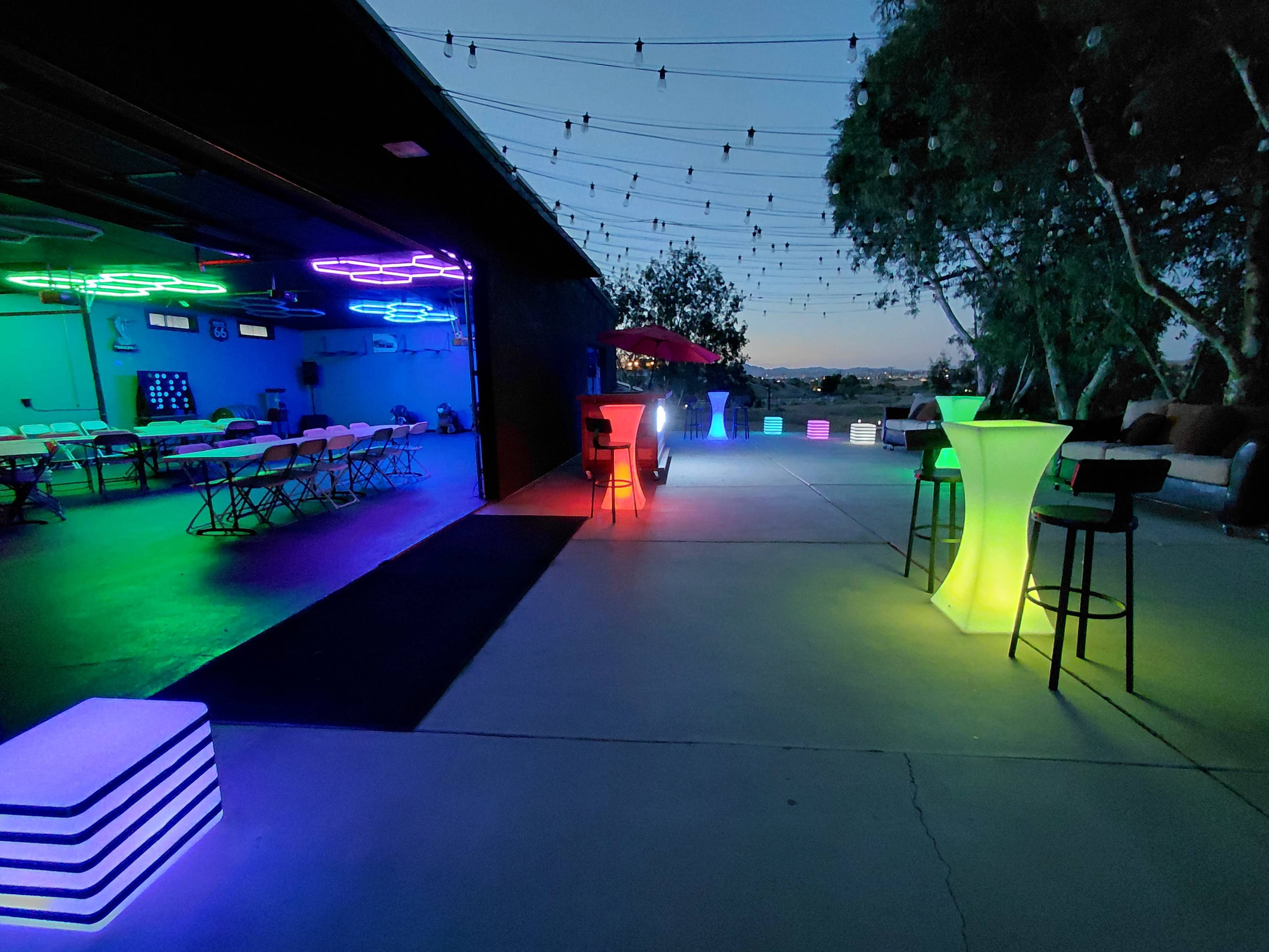 The scene features a brightly lit outdoor space with colorful LED furniture and string lights against a dusk sky.