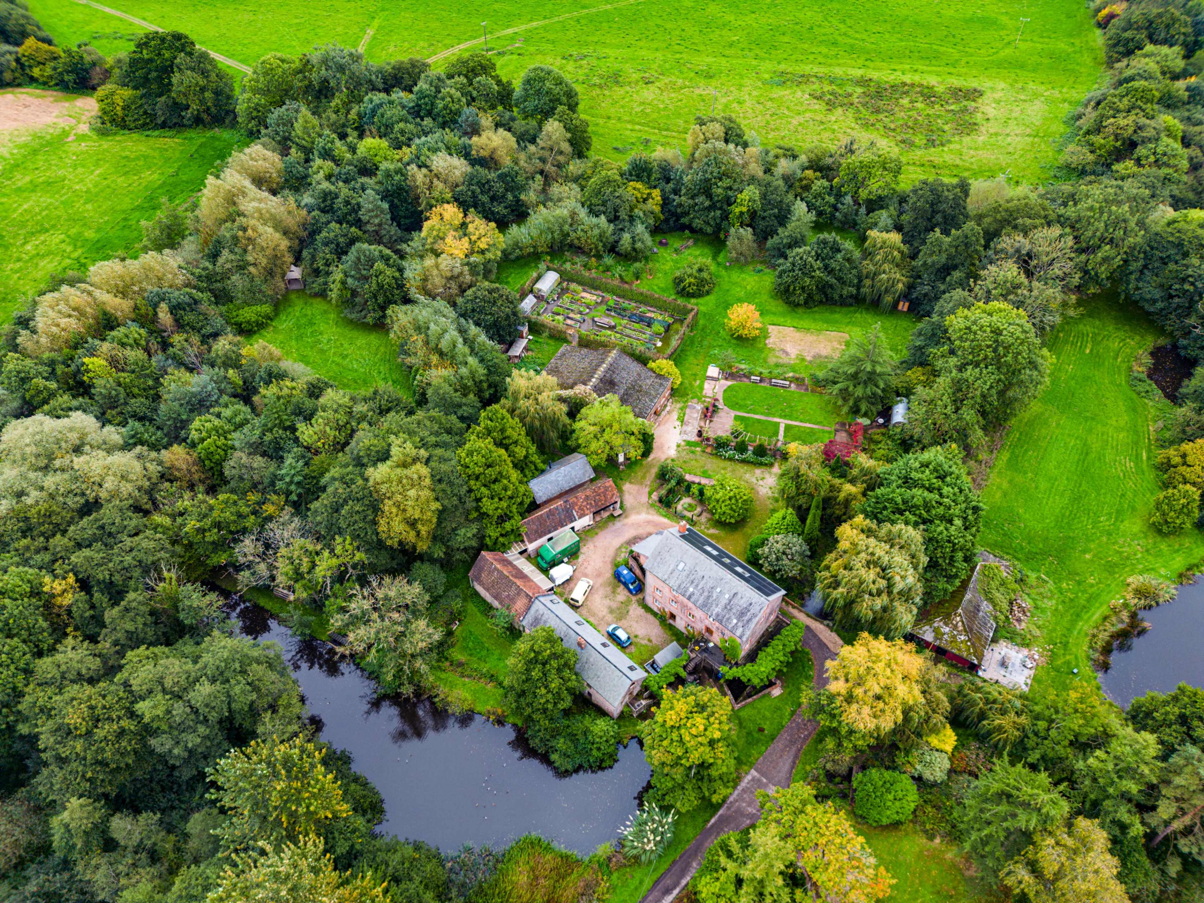 An aerial view shows a cluster of buildings and gardens surrounded by trees and water in a rural landscape.