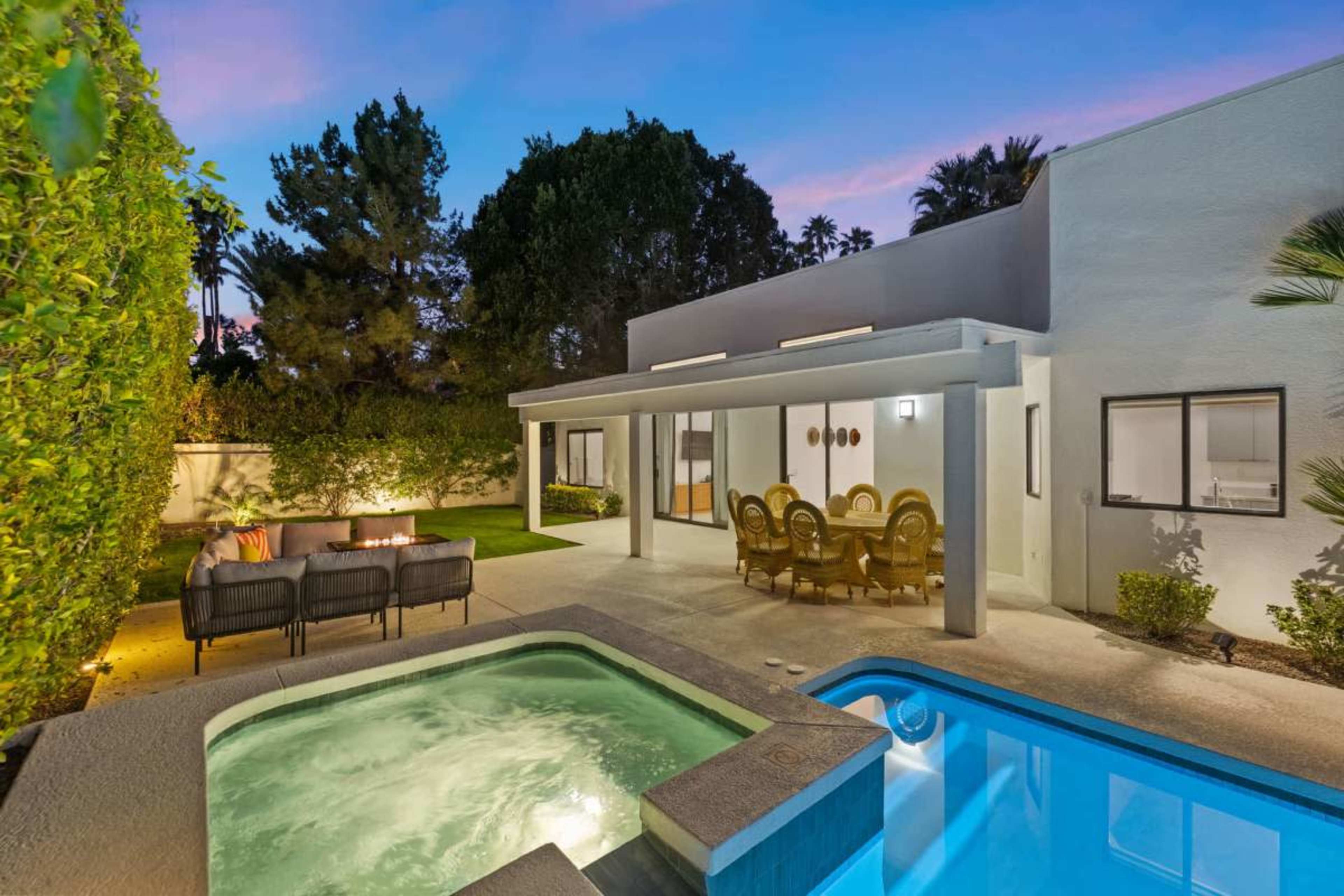The image shows a modern backyard with a swimming pool, a spa, a dining area under a covered patio, and lounge seating surrounded by greenery at dusk.