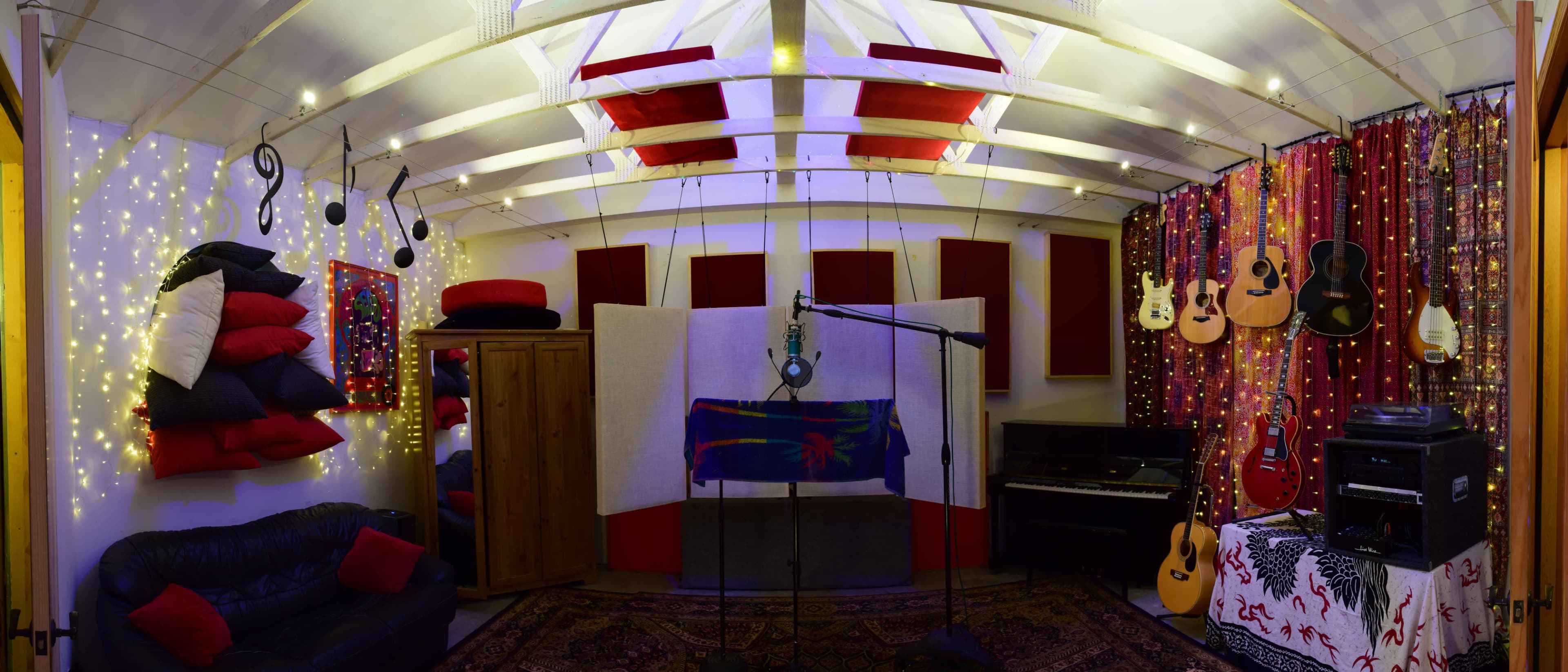 A music recording studio features a microphone on a stand surrounded by acoustic panels, guitars on the walls, and soft seating with decorative string lights.