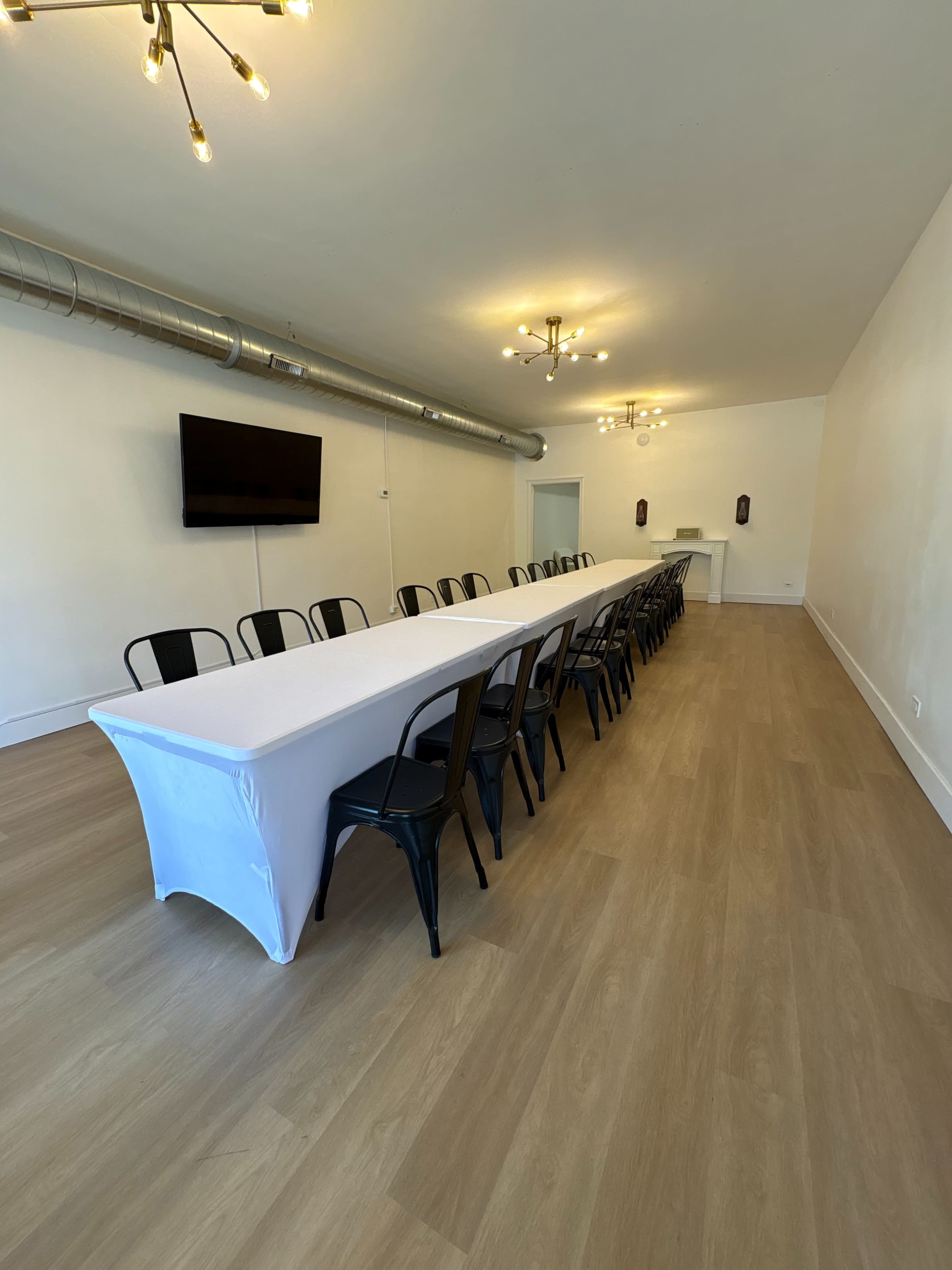 A long, rectangular table covered with a white tablecloth is set up in a spacious room with modern lighting fixtures and a television mounted on the wall.