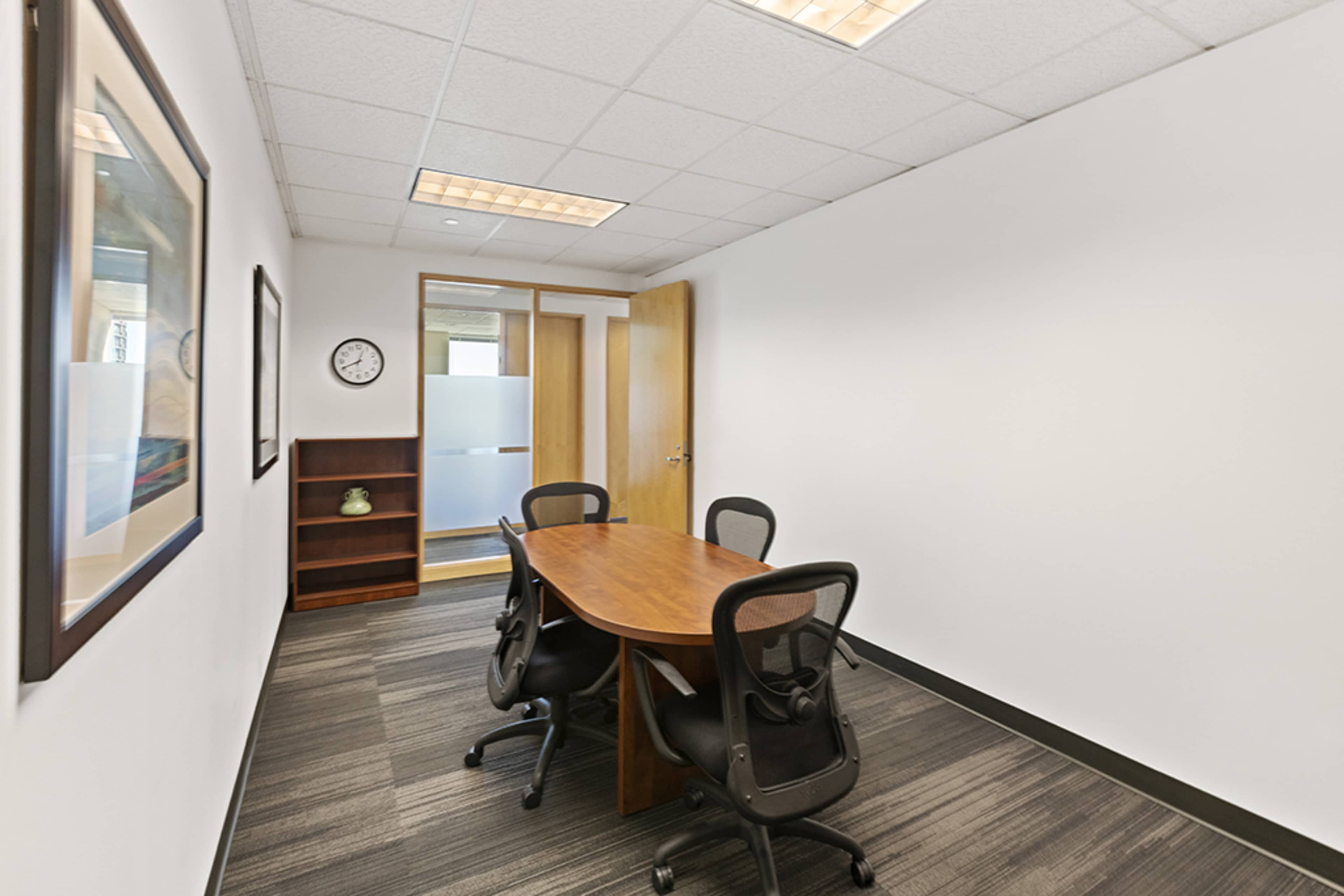A small conference room features a wooden table surrounded by four black chairs, with a clock and a bookcase on the walls.