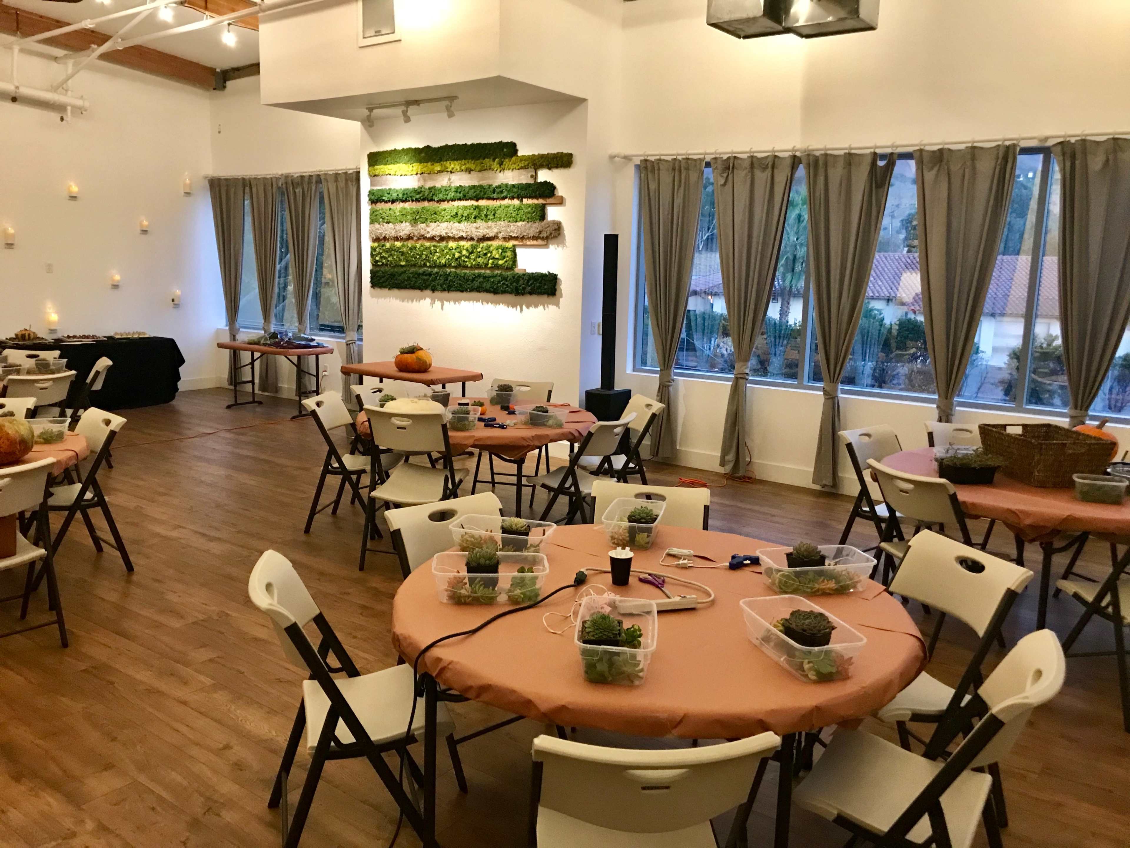 A spacious event room with round tables set for a gathering, featuring orange tablecloths and small centerpieces, along with large windows and greenery on the walls.