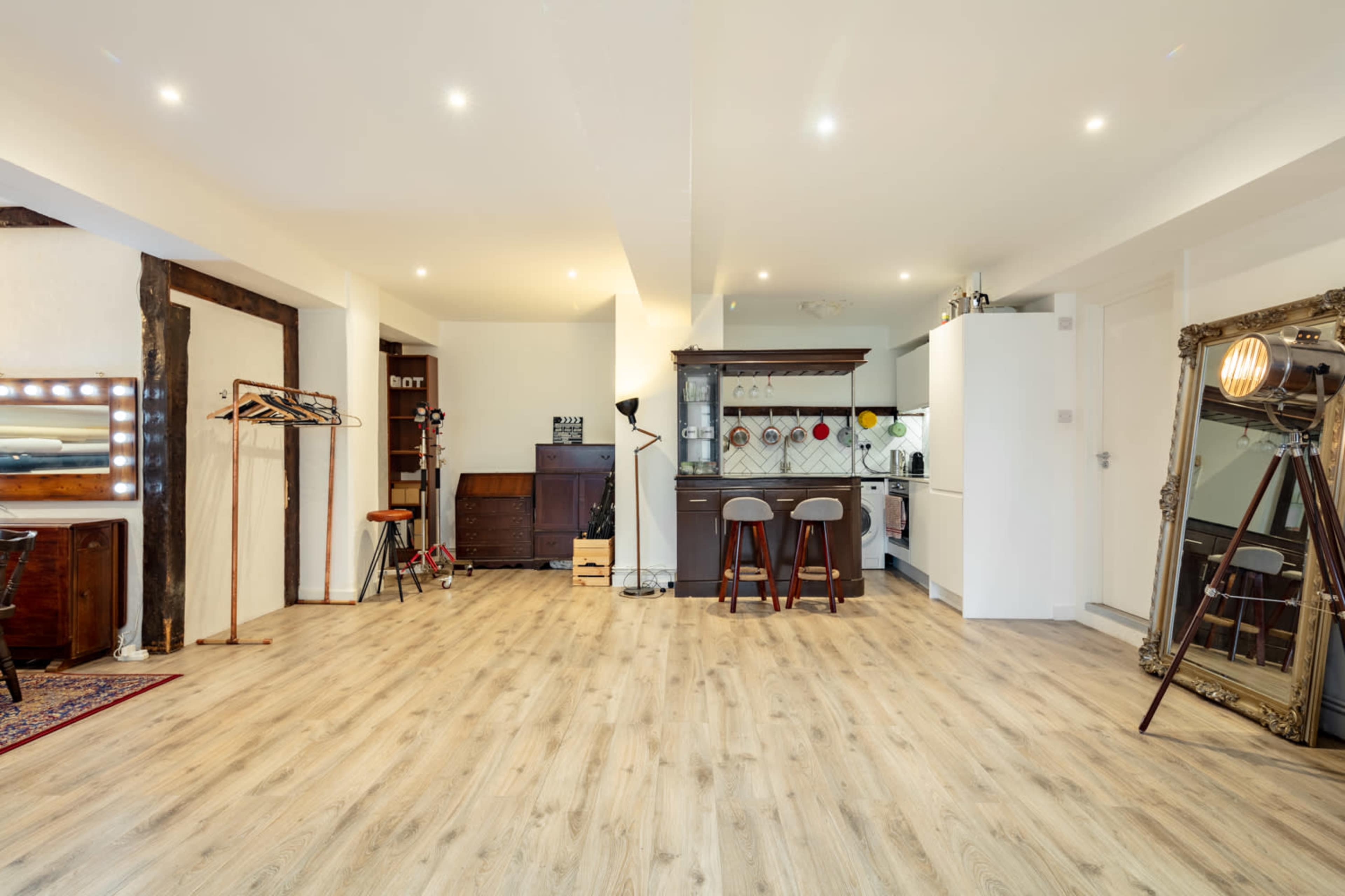 A spacious, well-lit room featuring wooden flooring, a bar area with two stools, and a combination of decorative furniture and mirrors.
