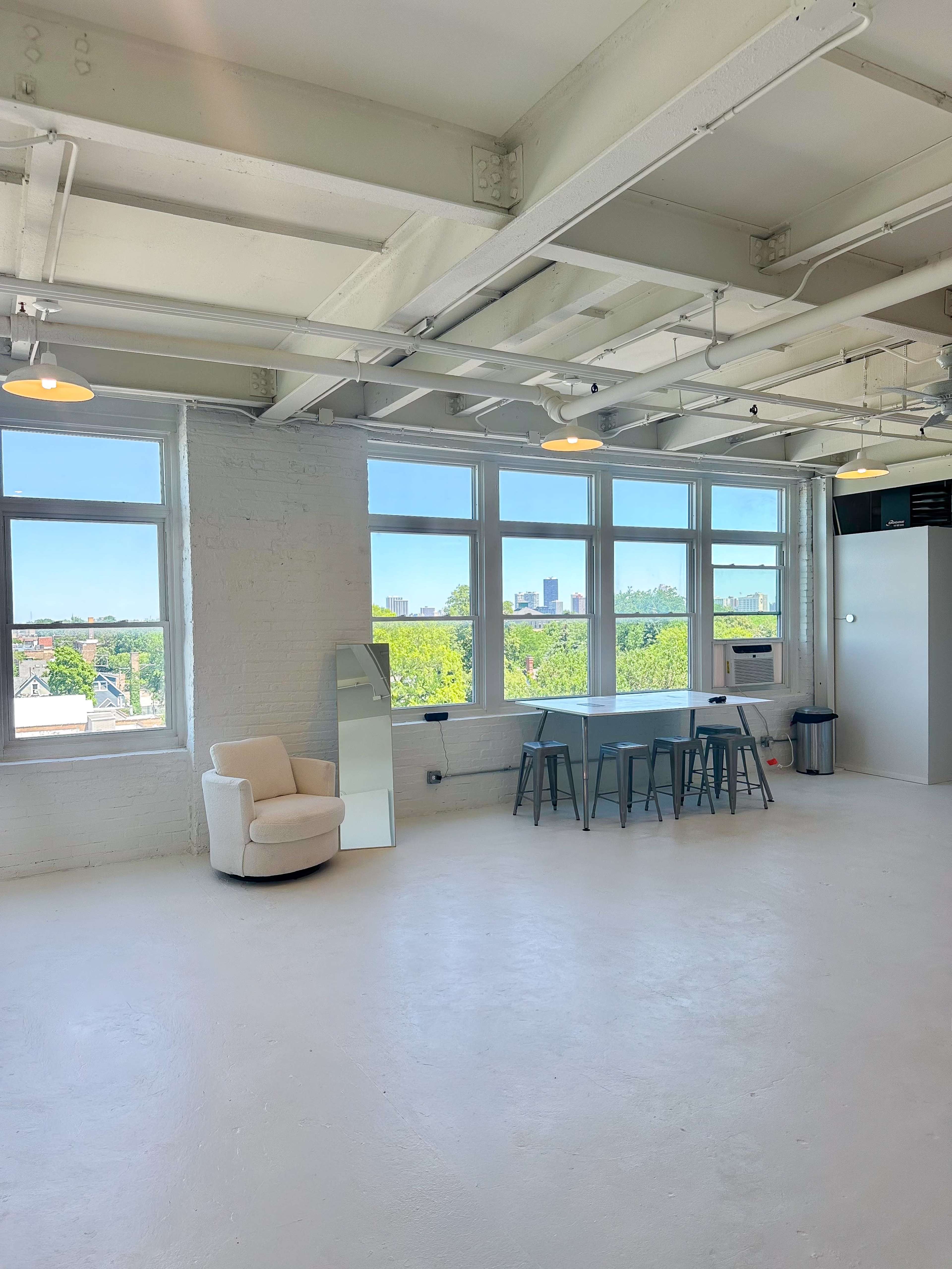 The image shows a spacious, brightly lit studio with large windows, a small table with stools, and a cozy chair against a white wall.