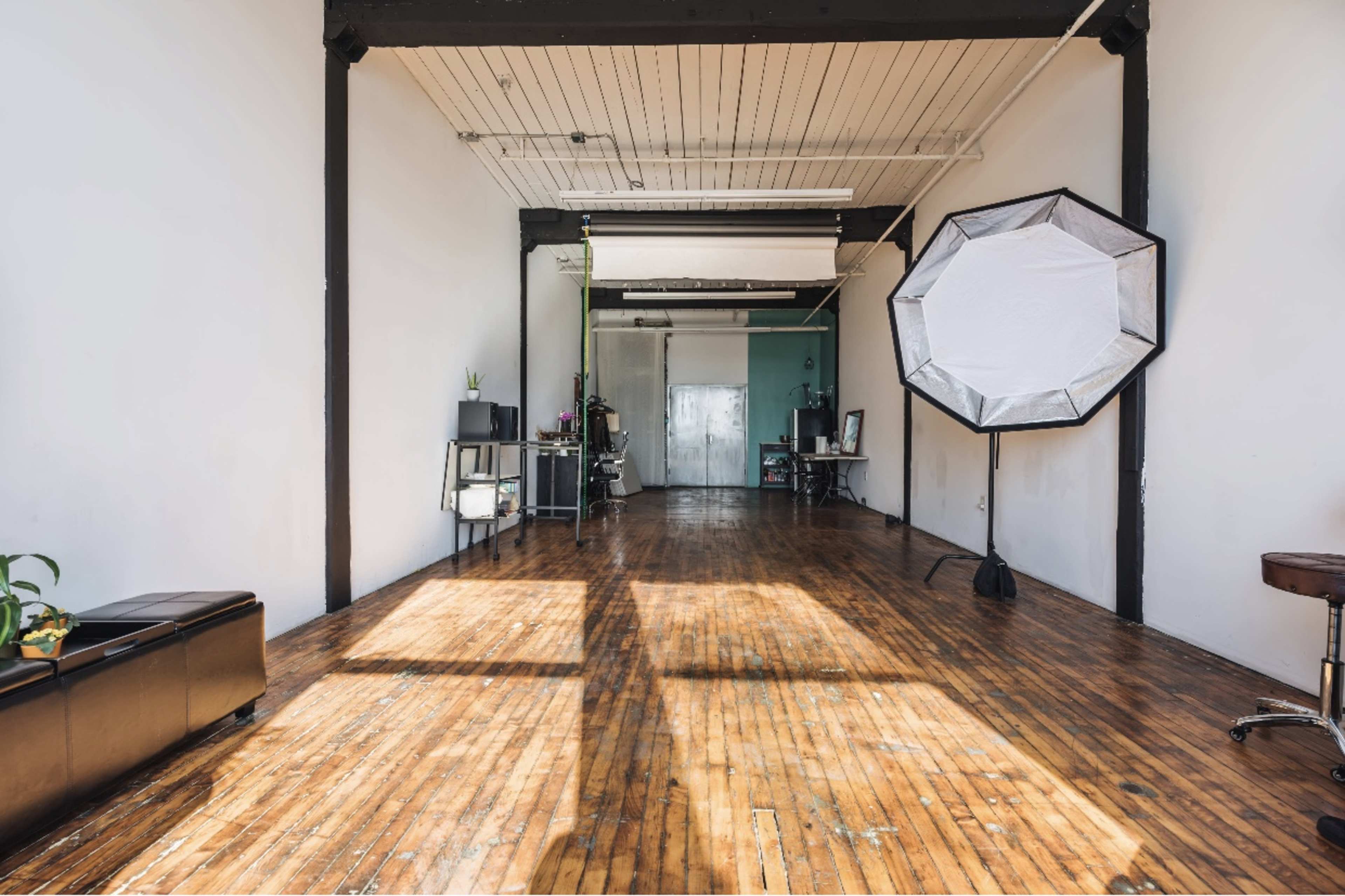 A spacious and minimalistic studio features wooden floors, large windows allowing natural light, and a large softbox setup in one corner.
