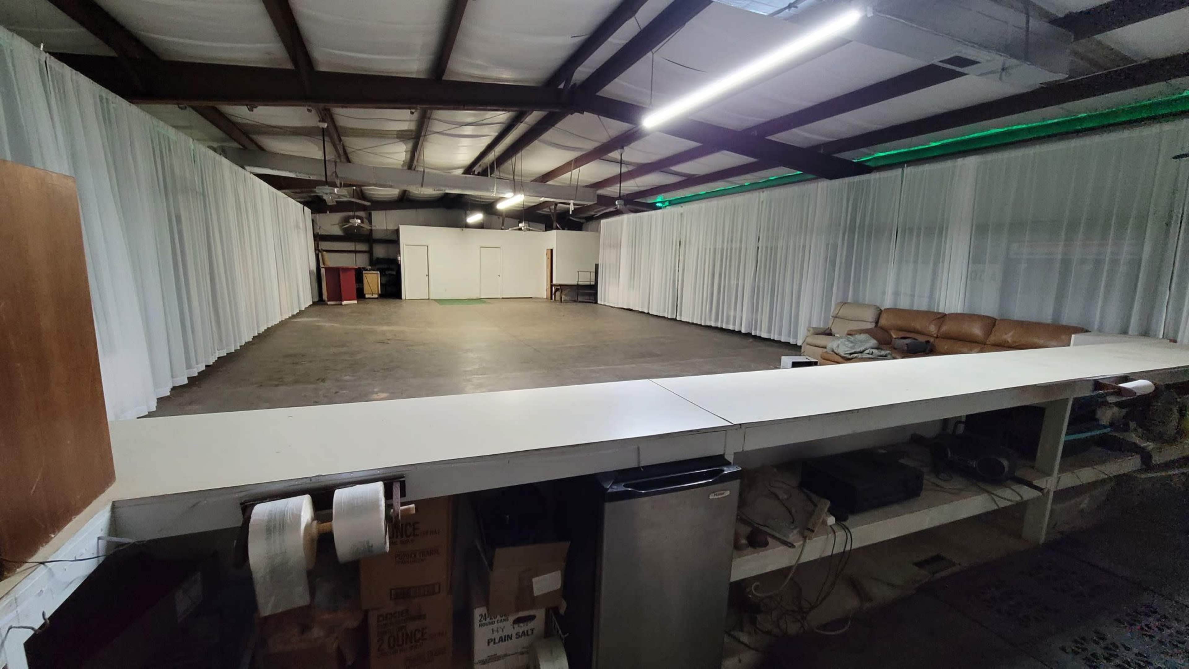 The image shows a large, empty room with a concrete floor, white drapes along the walls, and a brown couch in the corner next to a long counter with various appliances underneath.