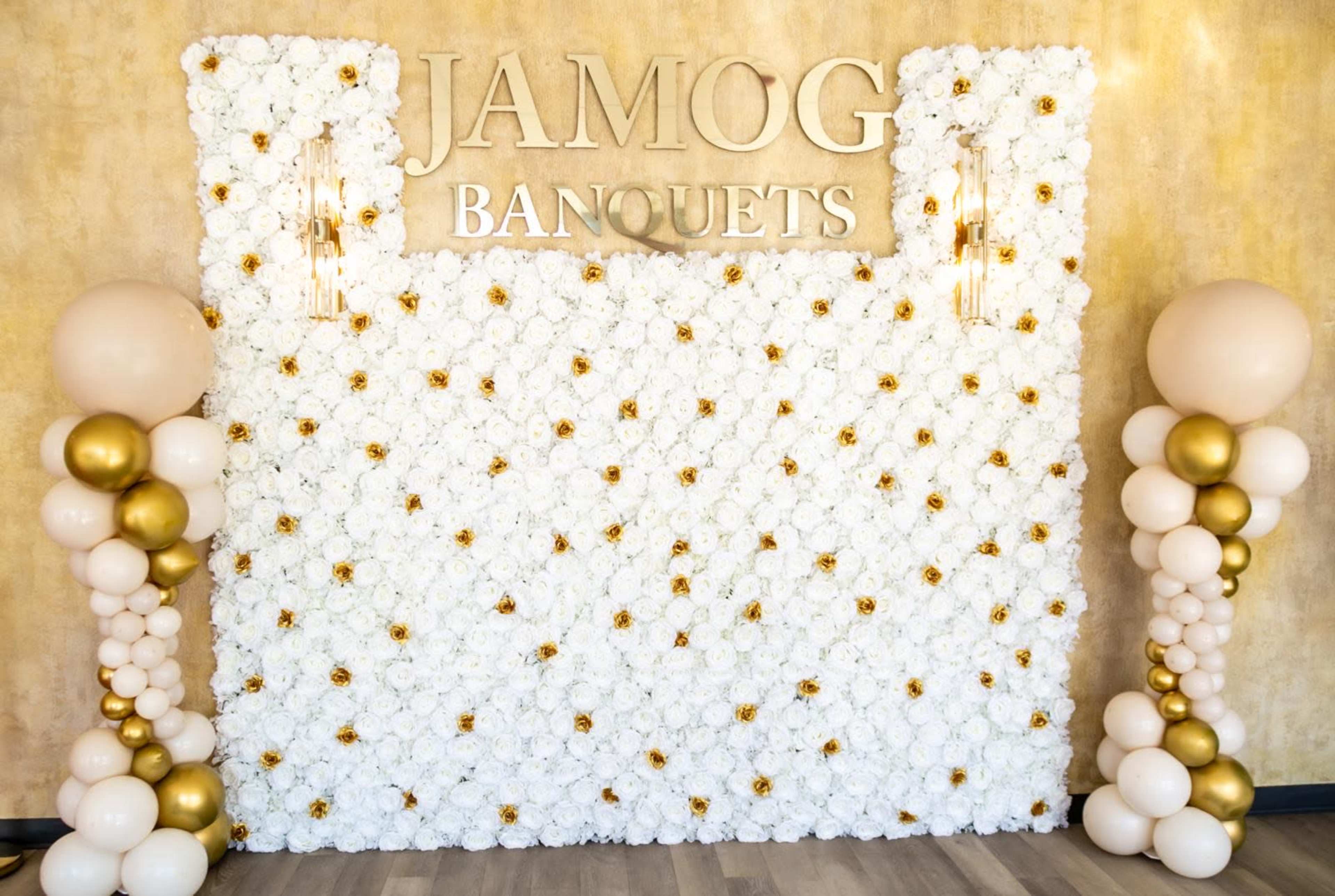 The image depicts a floral wall adorned with white flowers and decorative elements, featuring the text "JAMOG BANQUETS" in gold lettering, flanked by balloon columns in shades of white, gold, and light pink.