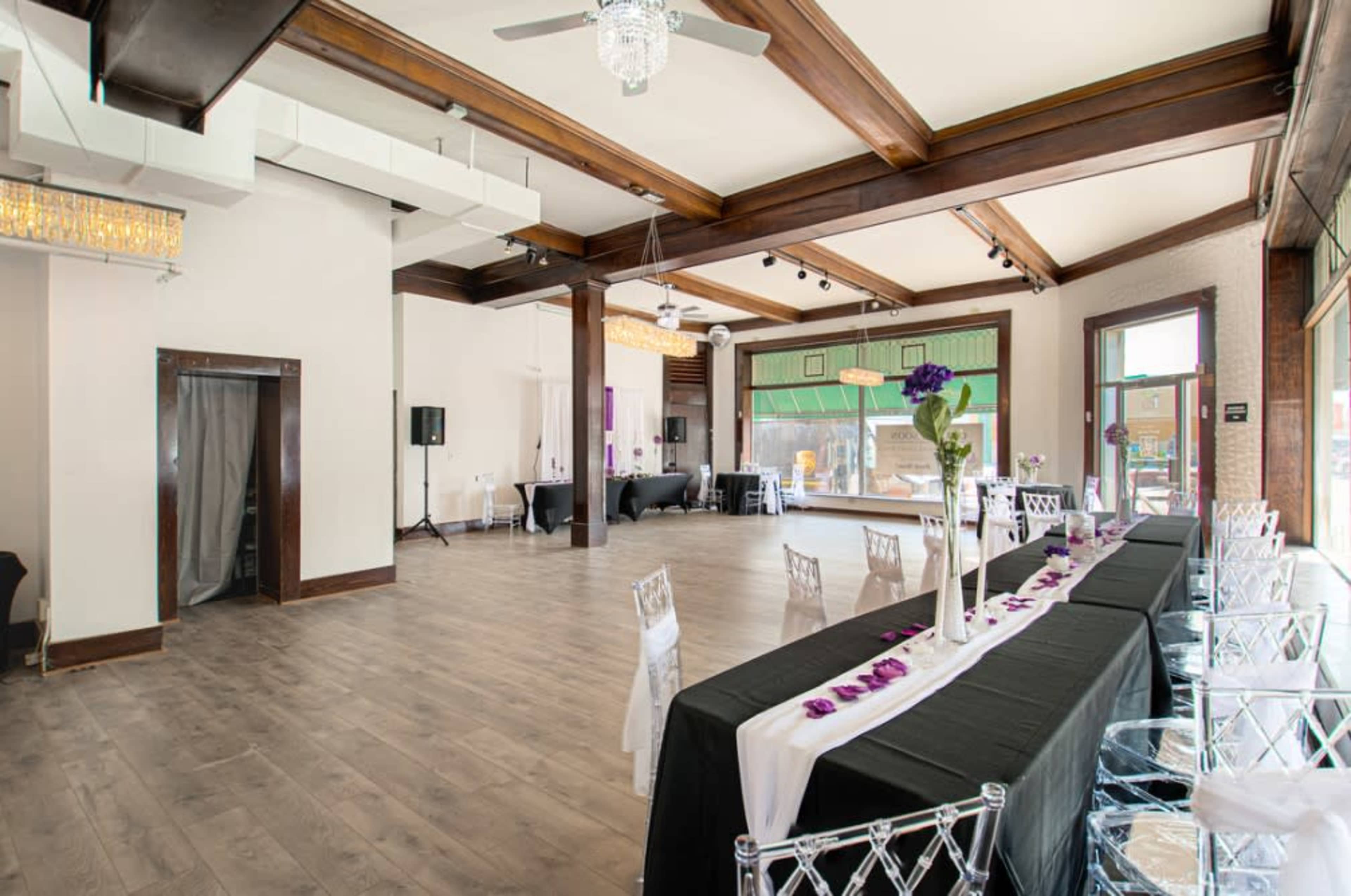 A spacious event venue with wooden beams, a light-colored wood floor, and a long table set with clear chairs and a black tablecloth adorned with purple petals.