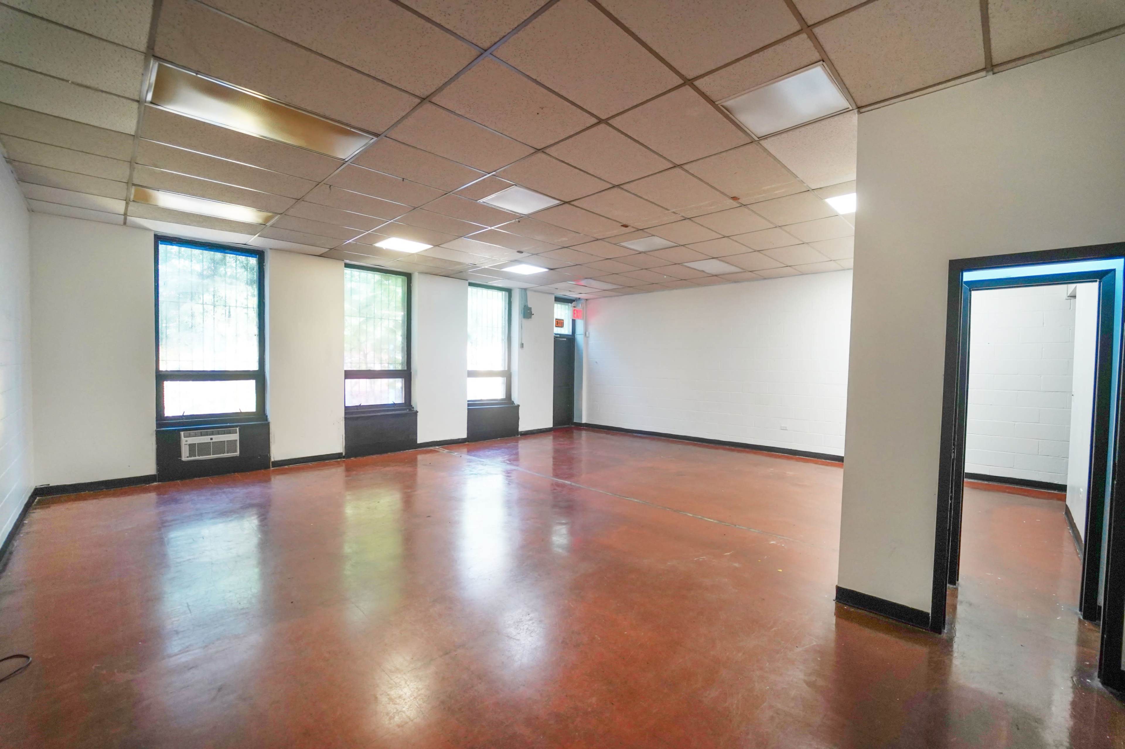 The image shows an empty room with a polished floor, featuring large windows on one side and two doorways leading to adjacent spaces.