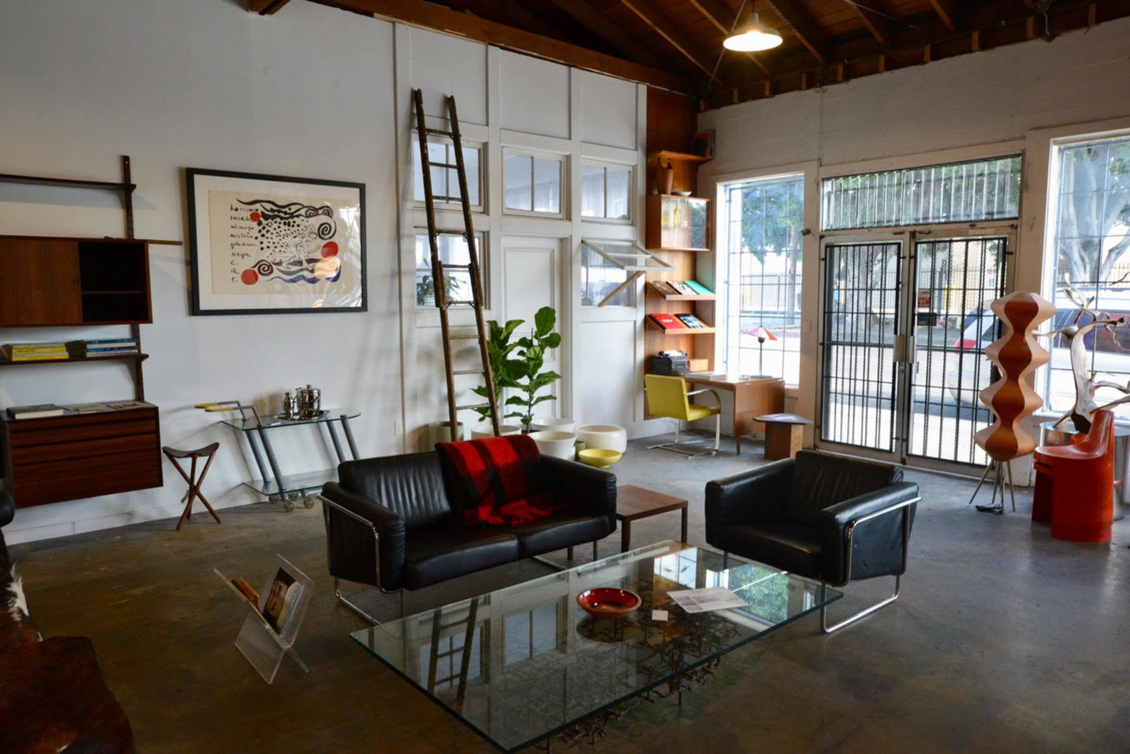 A minimalist interior with black leather sofas, a glass coffee table, and a wooden ladder against the wall, surrounded by various pieces of furniture and decor.