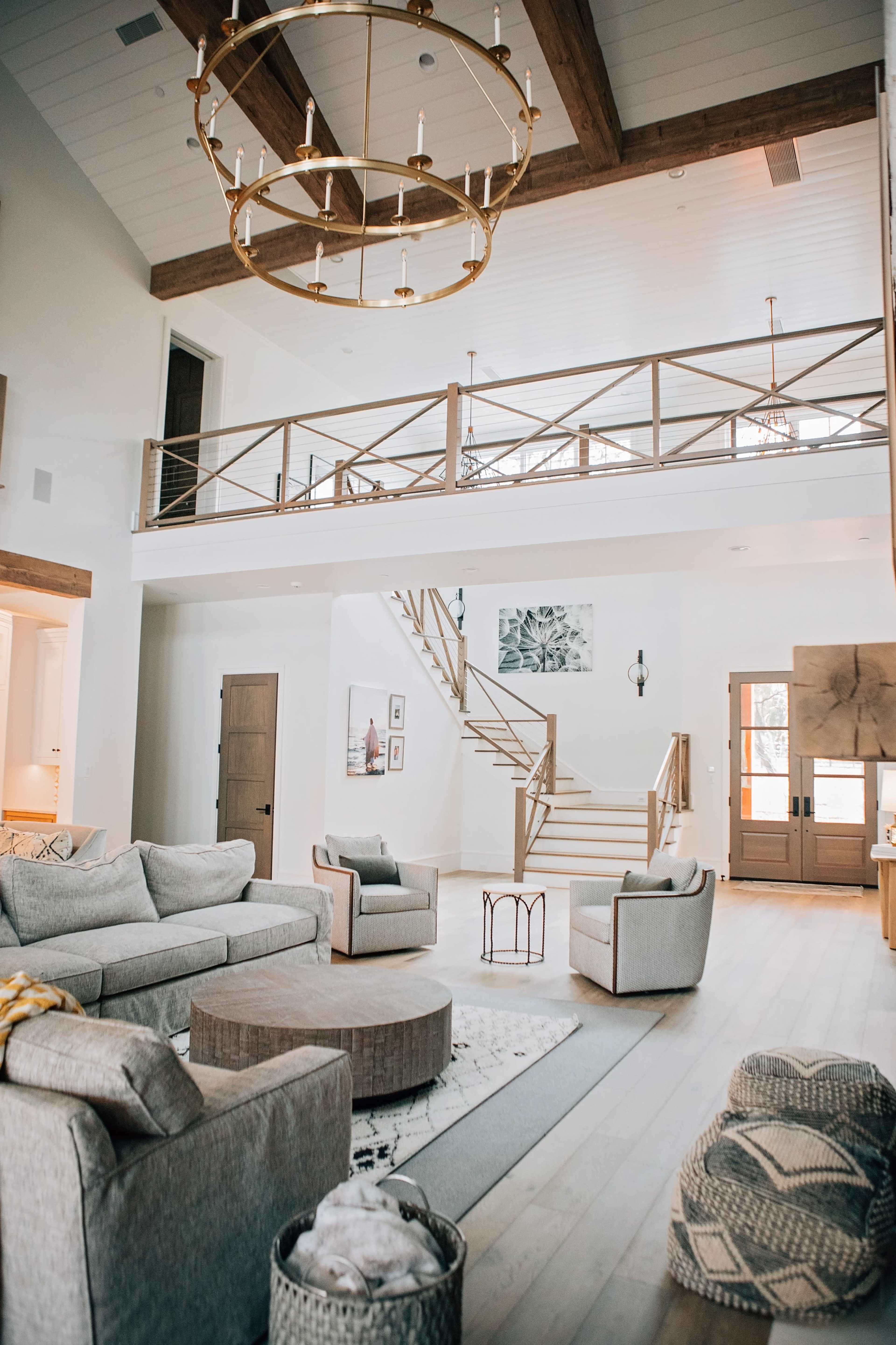 A spacious, modern living room with a high ceiling, featuring a staircase, multiple seating areas, and large windows.