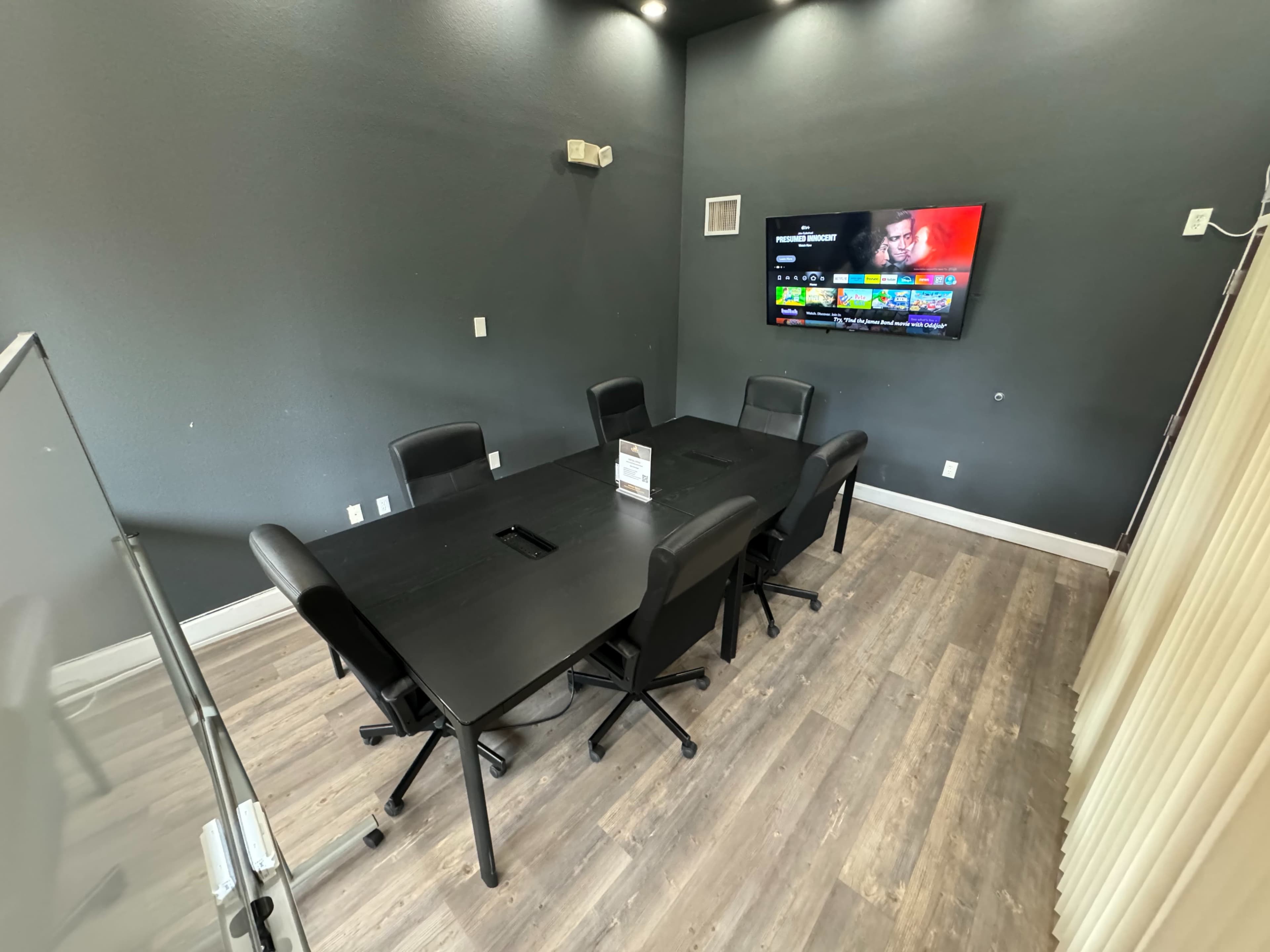 A conference room features a black rectangular table surrounded by six chairs and a wall-mounted TV.