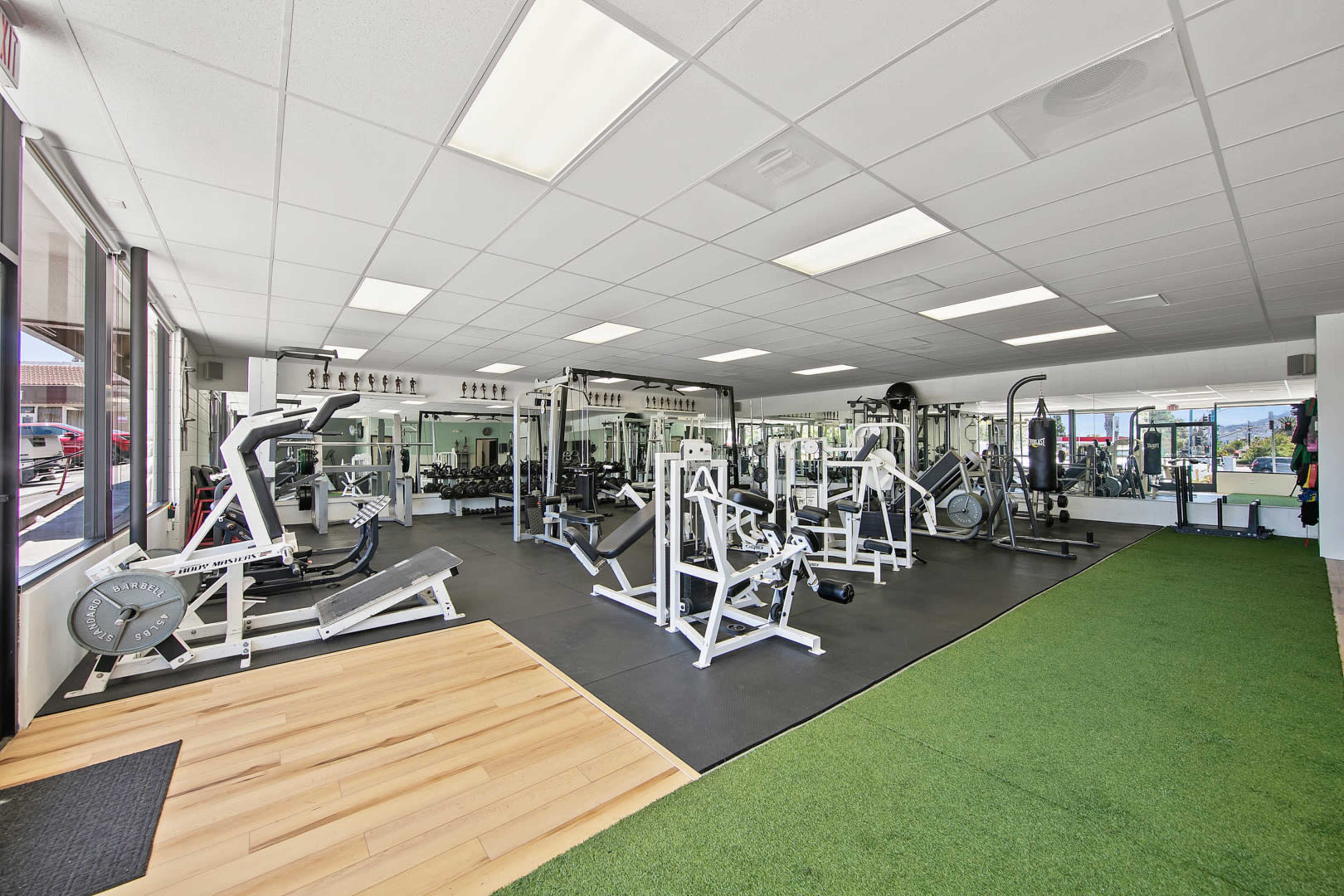 A spacious gym area with a variety of exercise equipment, including weight machines, cardio machines, and free weights, all set on a combination of rubber flooring and artificial turf.