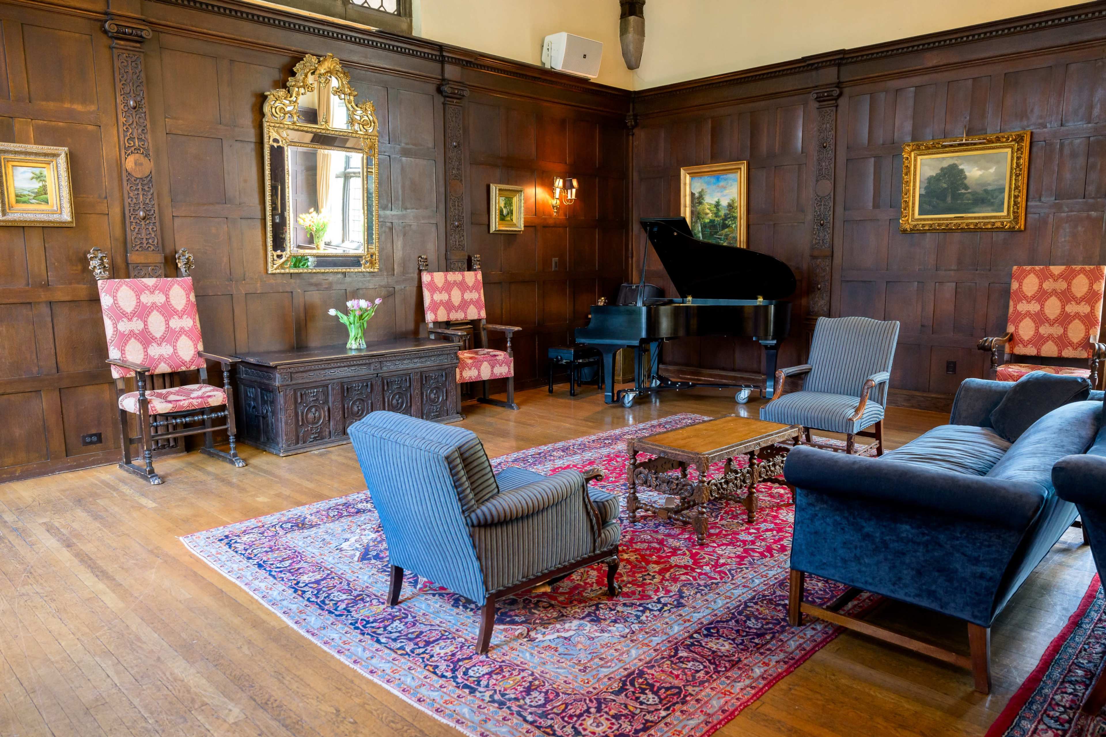 A wooden-paneled room featuring a grand piano, ornate furniture, and a large mirror, with a decorative rug on the floor.